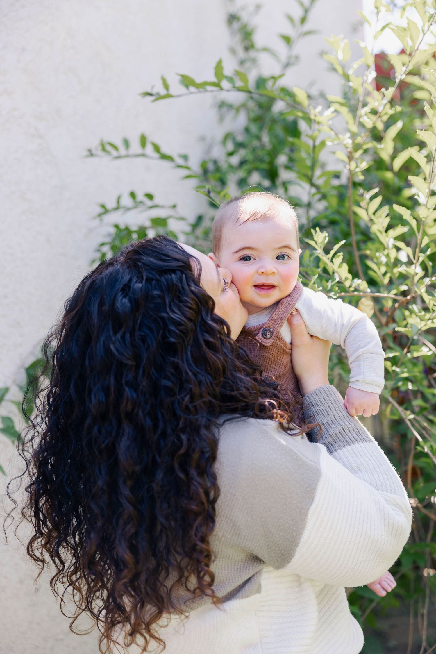A person with long dark curly hair holds a smiling baby outdoors against a light wall with green foliage in the background.