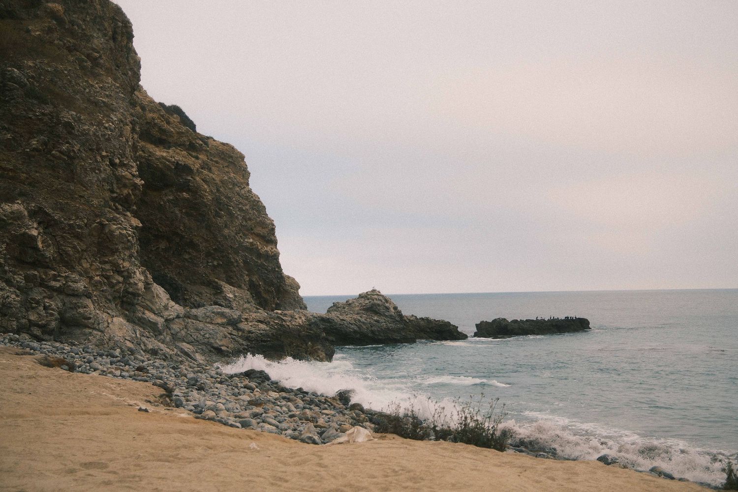 Rocky coastal cliffs meet a sandy beach as waves crash against the shore under an overcast sky.