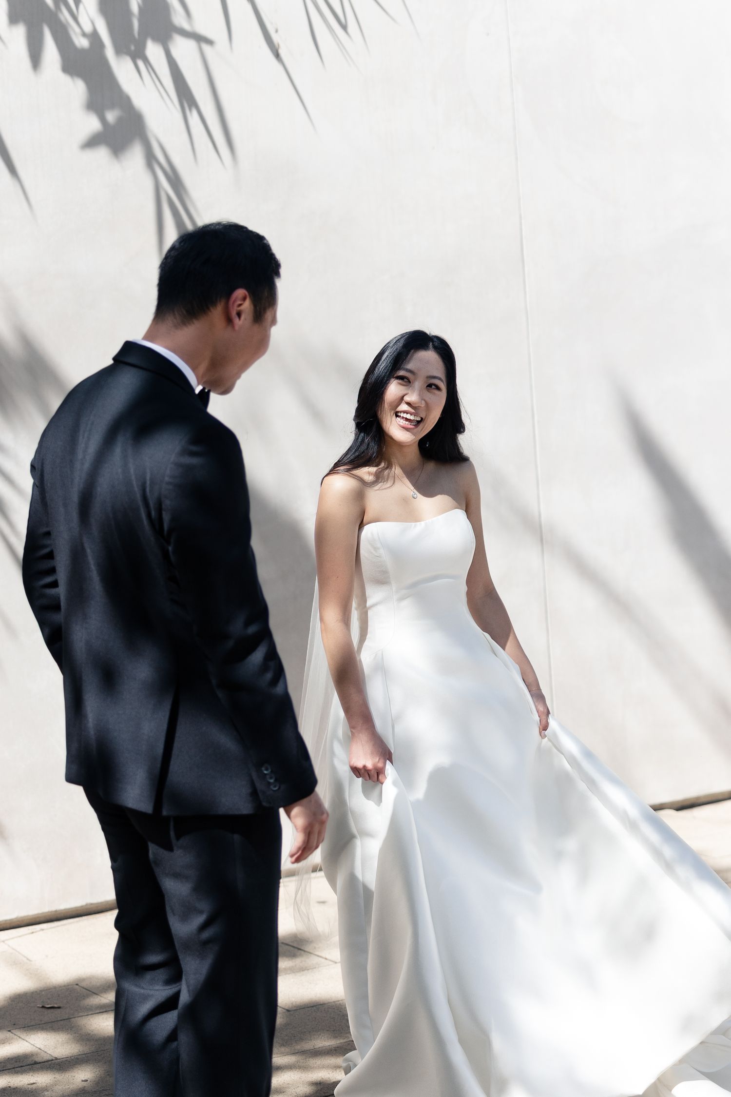 A smiling bride in a white strapless gown holds her dress while facing a groom in a black suit against a sunlit wall.