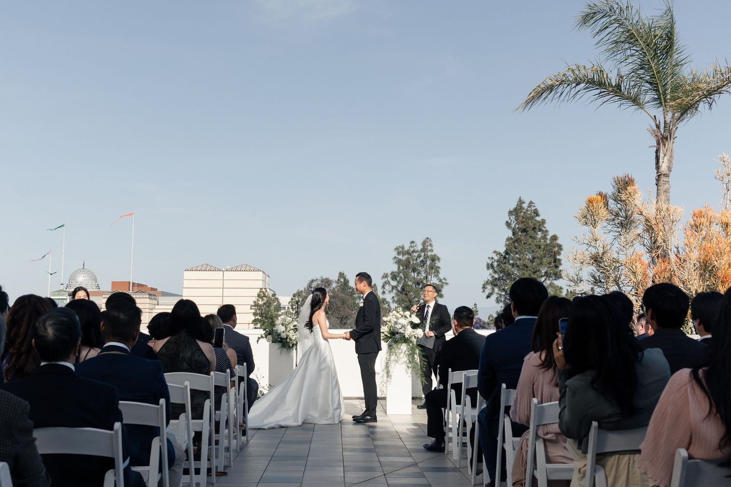 Outdoor rooftop wedding ceremony with bride and groom at altar, white chairs, guests seated, palm trees, and blue sky backdrop.