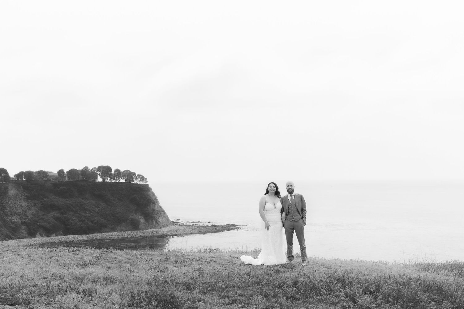 Black and white photo of a bride and groom standing on a coastal cliff with ocean views and a lighthouse in the background.