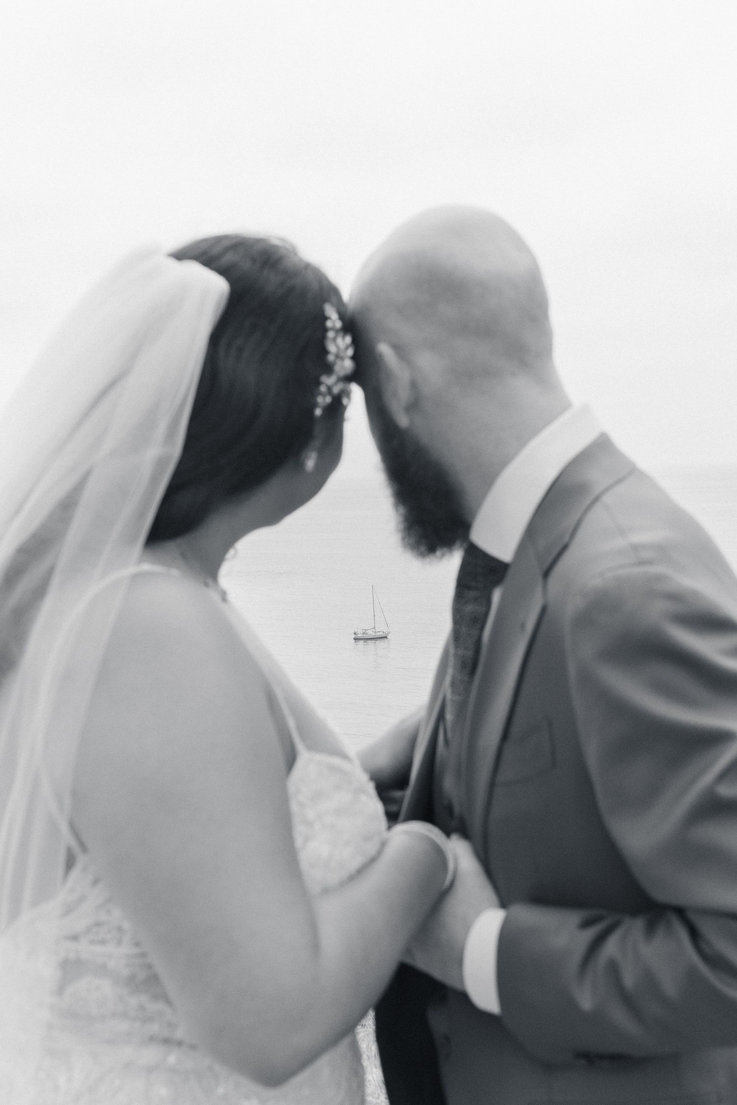 Black and white photo of a bride and groom holding hands, leaning foreheads together near a serene waterfront.