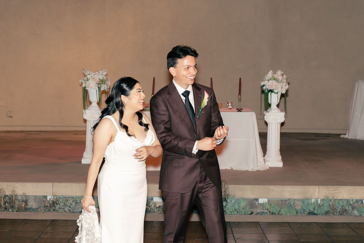 A bride in a white lace gown and groom in a dark suit walk together at their wedding reception venue.