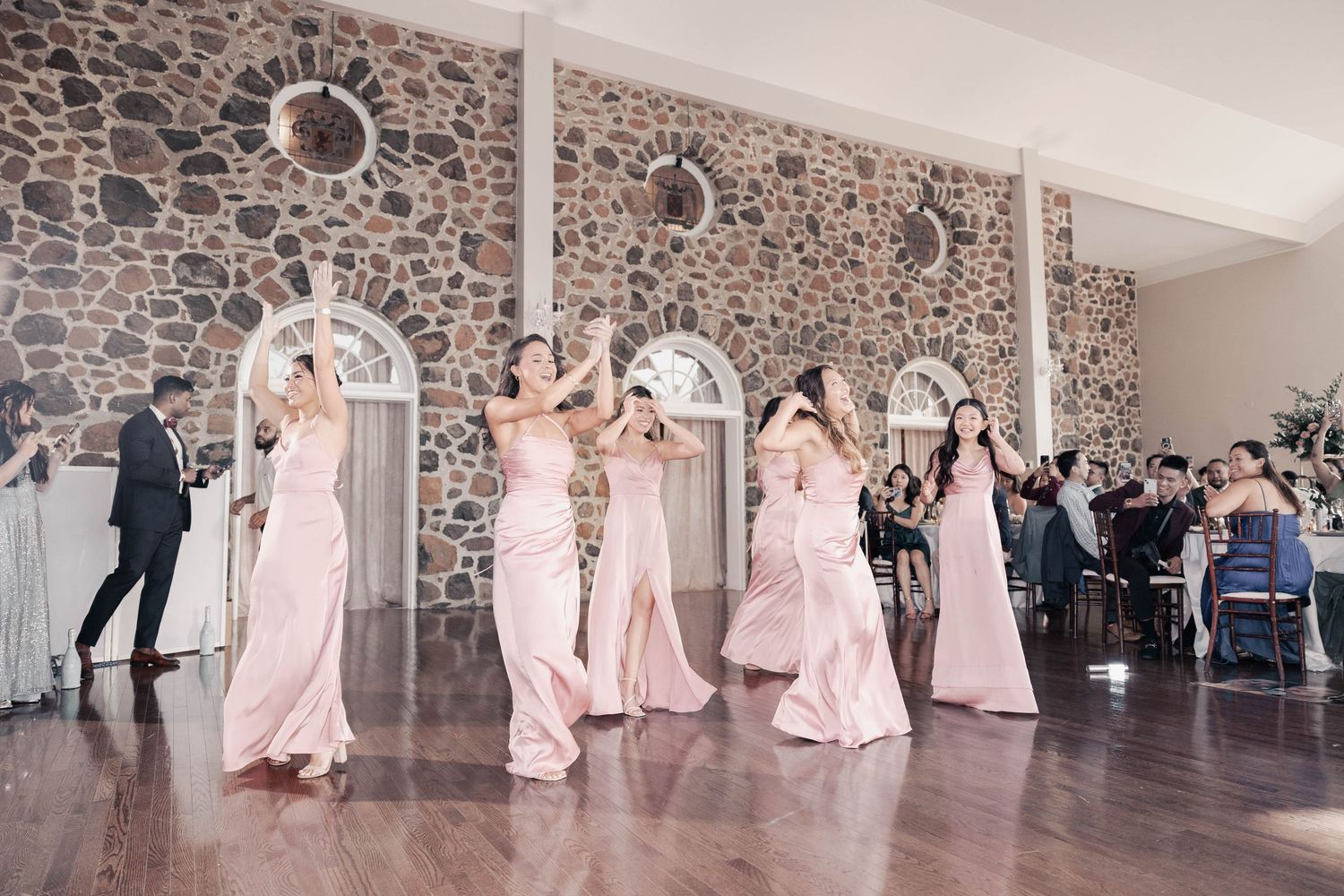Wedding party dancing in a rustic stone-walled reception venue with pink bridesmaid dresses.