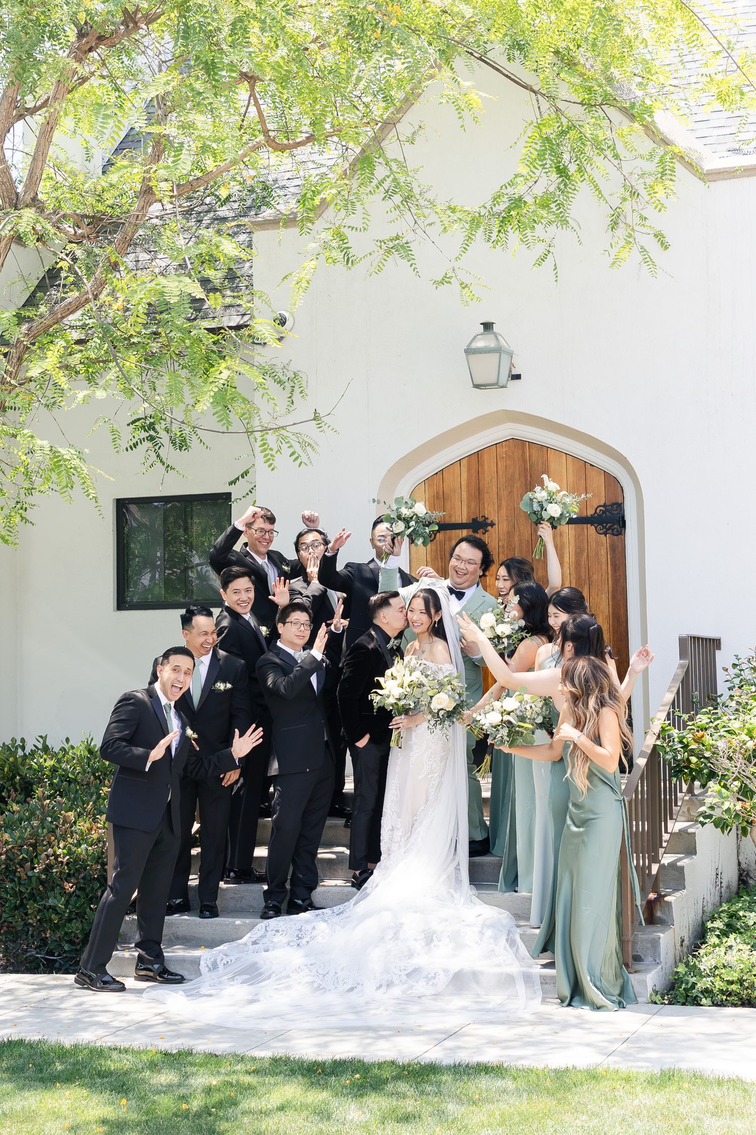 Wedding party celebrates outside a white chapel with wooden doors, bridesmaids in sage green gowns and groomsmen in black suits.