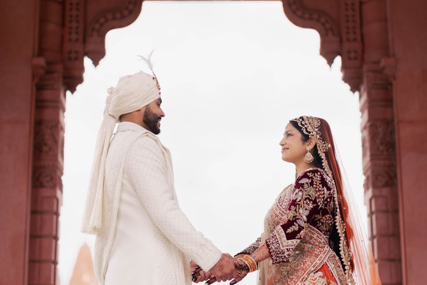 A couple in traditional Indian wedding attire stands in an ornate archway with stone columns.