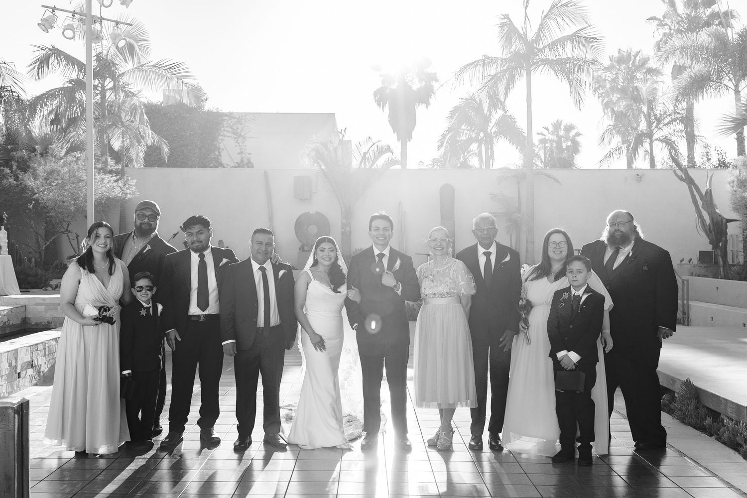 Black and white wedding group photo with bride and groom surrounded by family outdoors with palm trees.