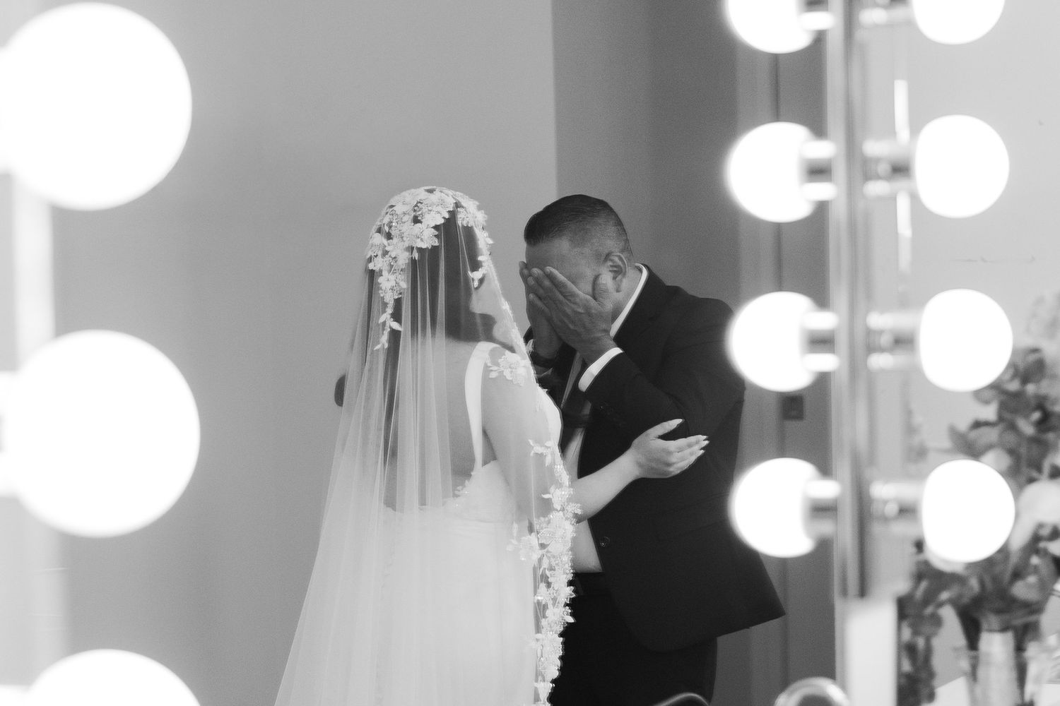 Black and white wedding photo of bride and groom sharing an intimate moment near a vanity mirror with glowing bulbs.