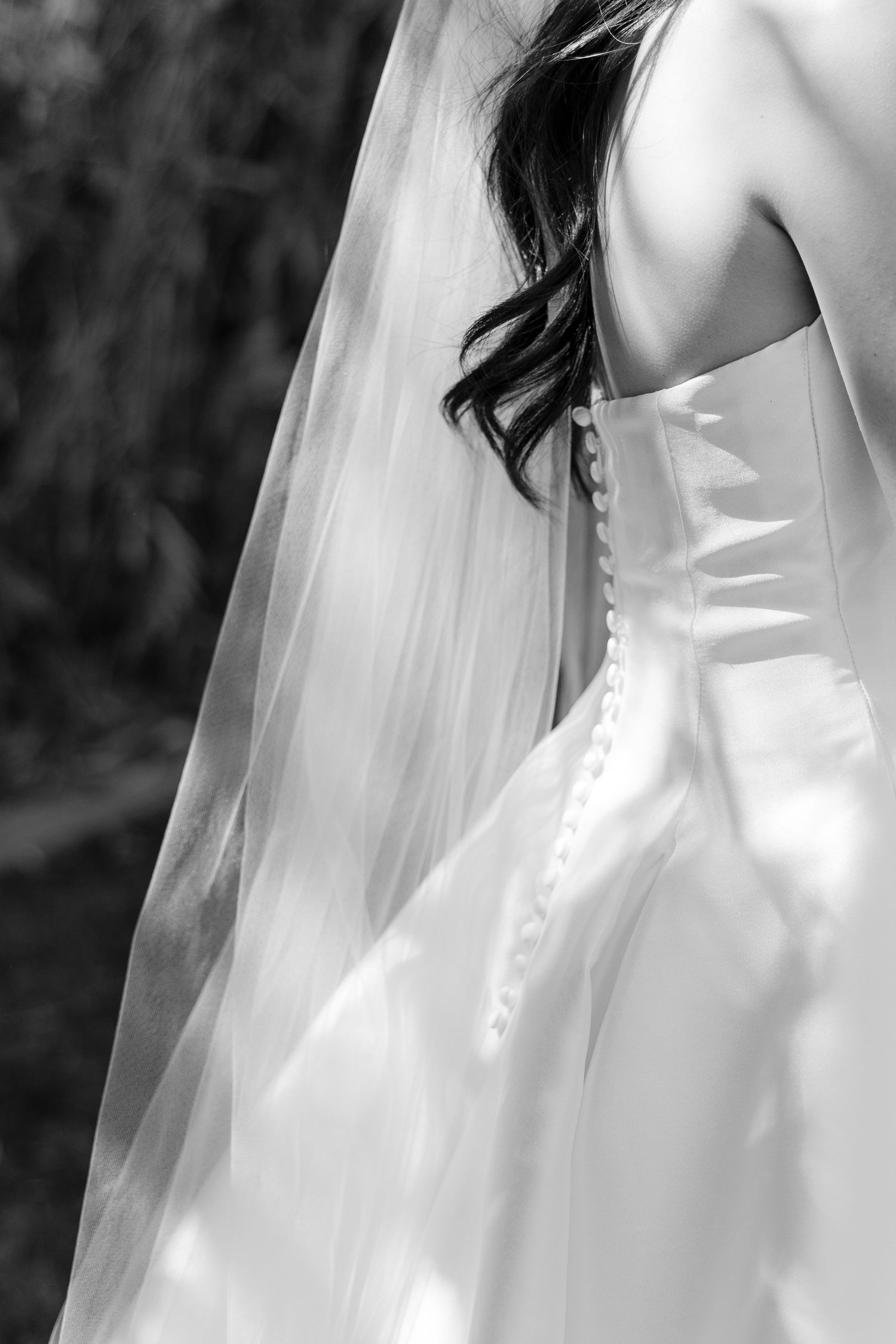 Black and white photo of a bride in a satin corset wedding gown with pearl buttons and flowing veil.