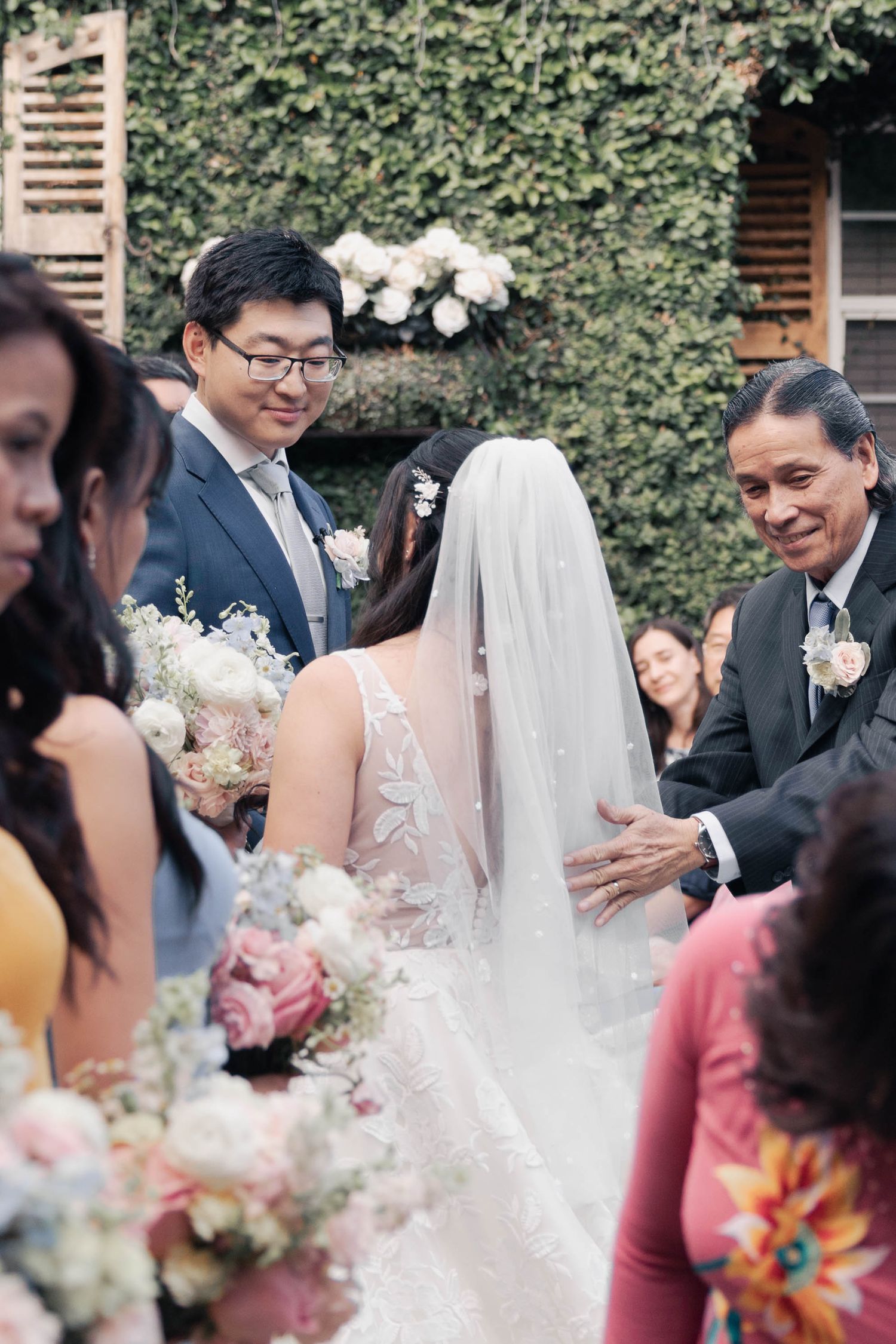 Wedding guests gather outdoors at a garden ceremony with brick walls and ivy creating an elegant backdrop.