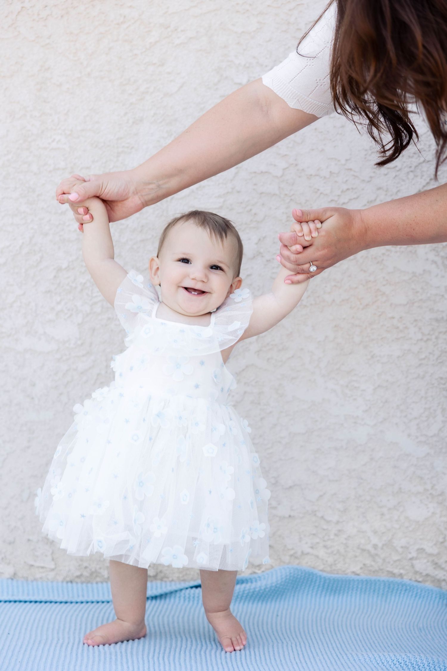 A baby in a white tulle dress takes first steps on a blue mat while supported by adult hands.