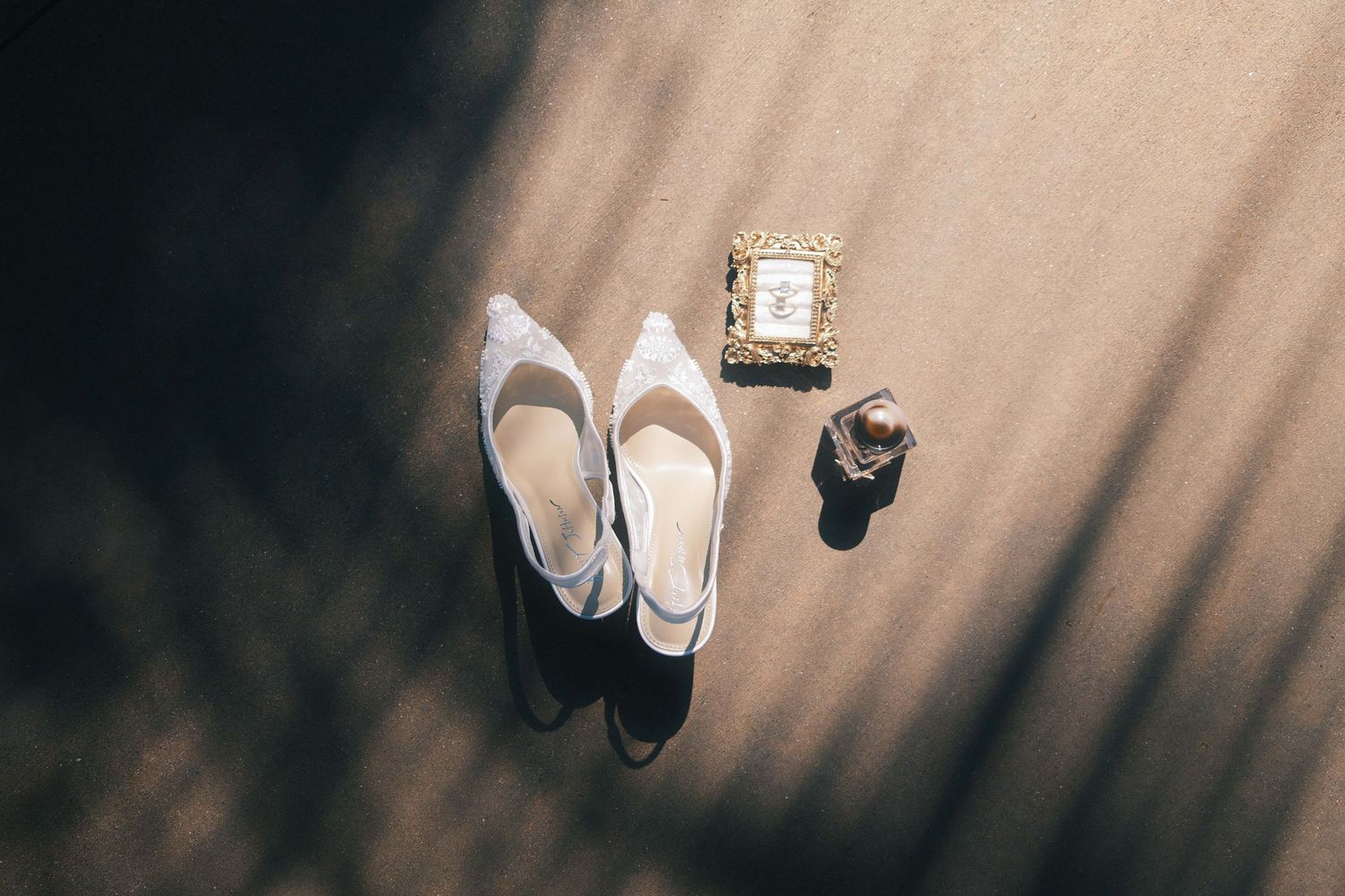 White bridal slingback heels, gold picture frame, and ring displayed on sandy surface with dramatic shadows.