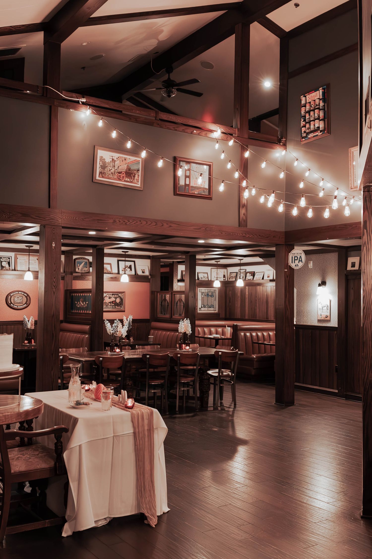 A cozy restaurant interior with wood beams, string lights, and rustic decor features tables set with white tablecloths.