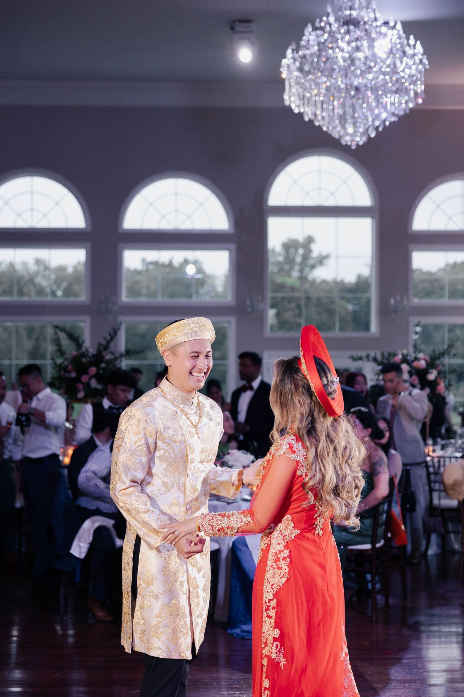 Guests in traditional Asian formal wear dancing beneath crystal chandeliers at an elegant venue for their wedding.
