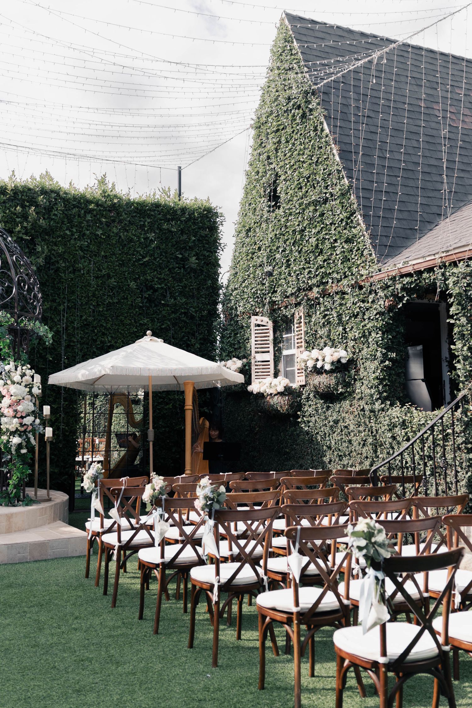 Outdoor wedding ceremony setup with wooden cross-back chairs arranged on grass lawn with ivy-covered walls and white umbrellas.