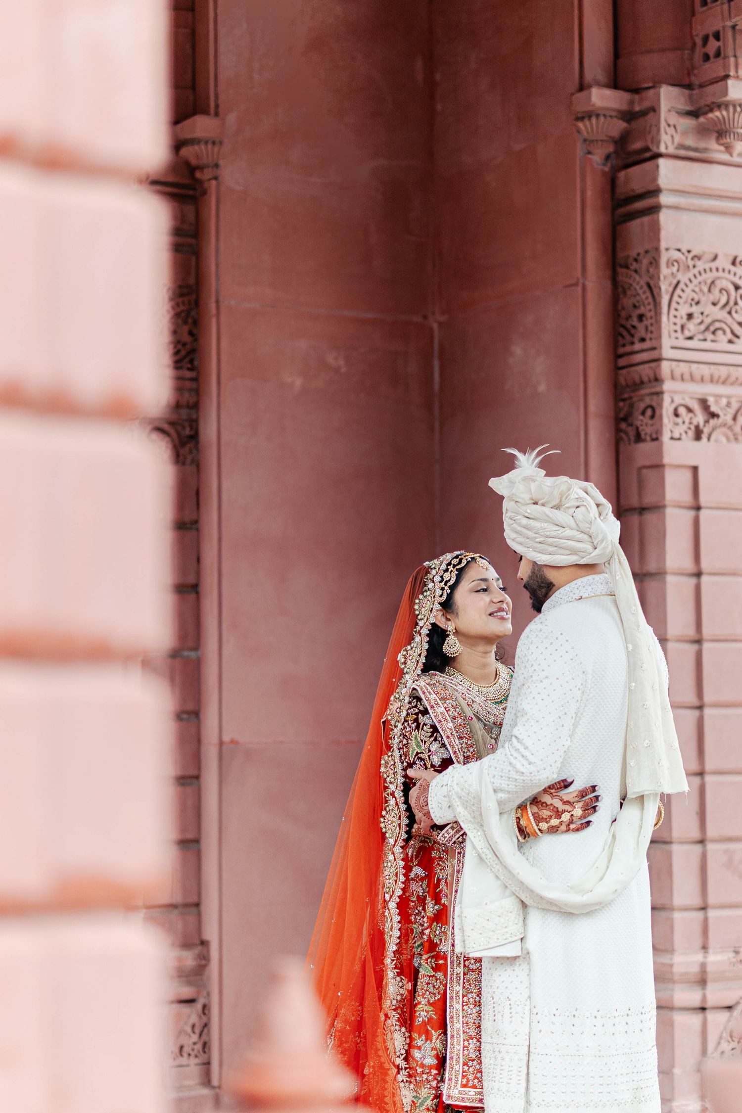A couple in traditional South Asian wedding attire embrace in front of ornate architectural details in warm tones.
