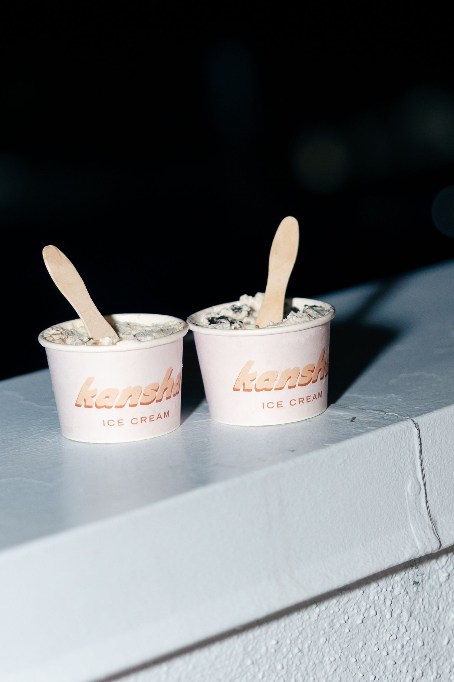 Two Kansha ice cream cups with wooden spoons on a white ledge against a dark background.
