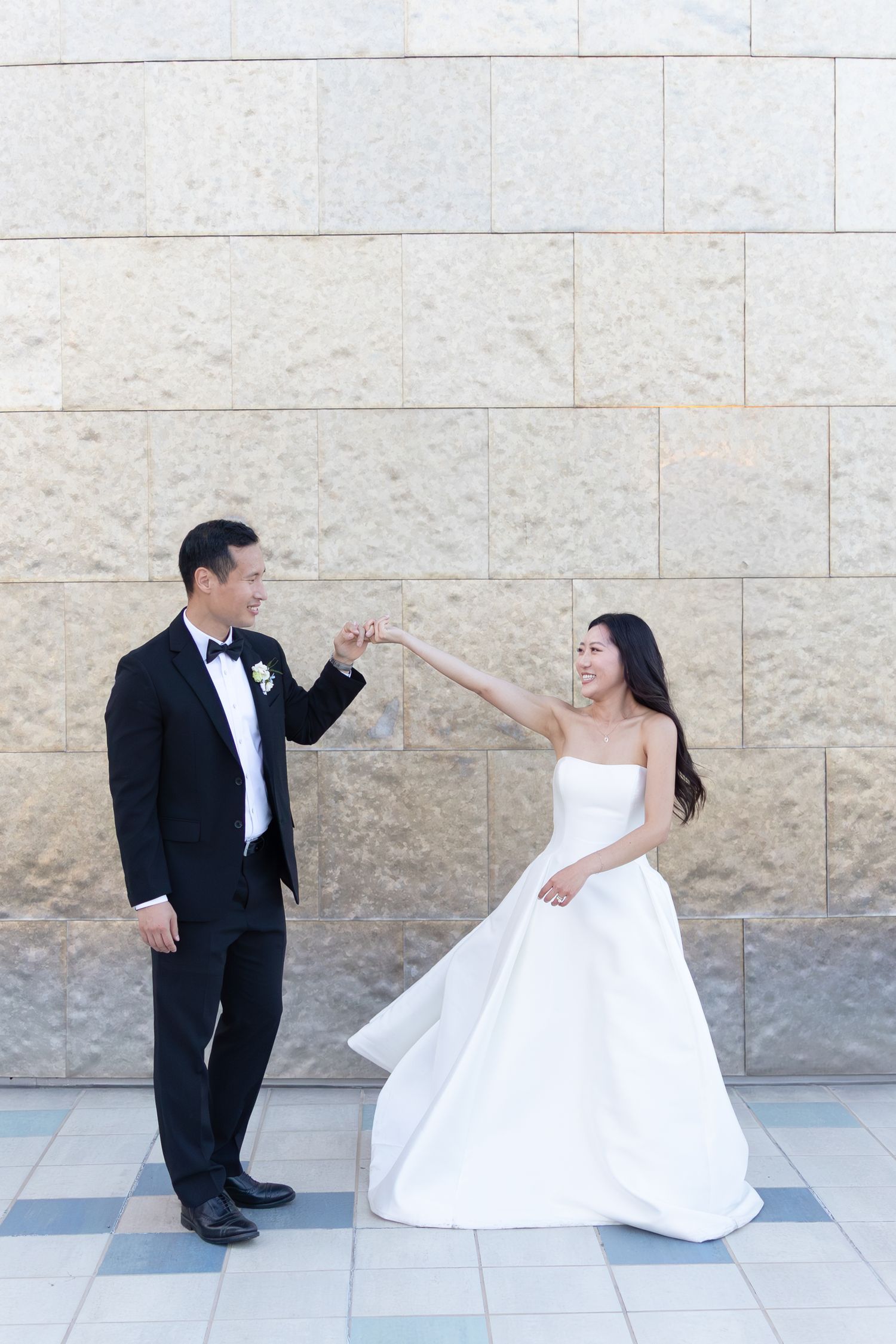 Bride and groom dancing joyfully in front of a stone wall, wearing a white gown and black tuxedo.