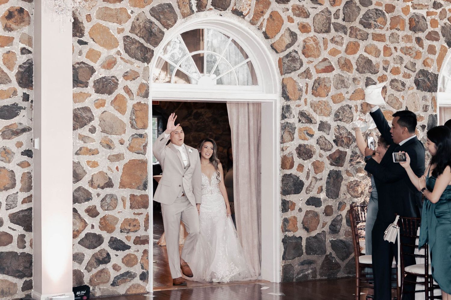 Wedding reception moment captured against rustic stone wall with arched windows.