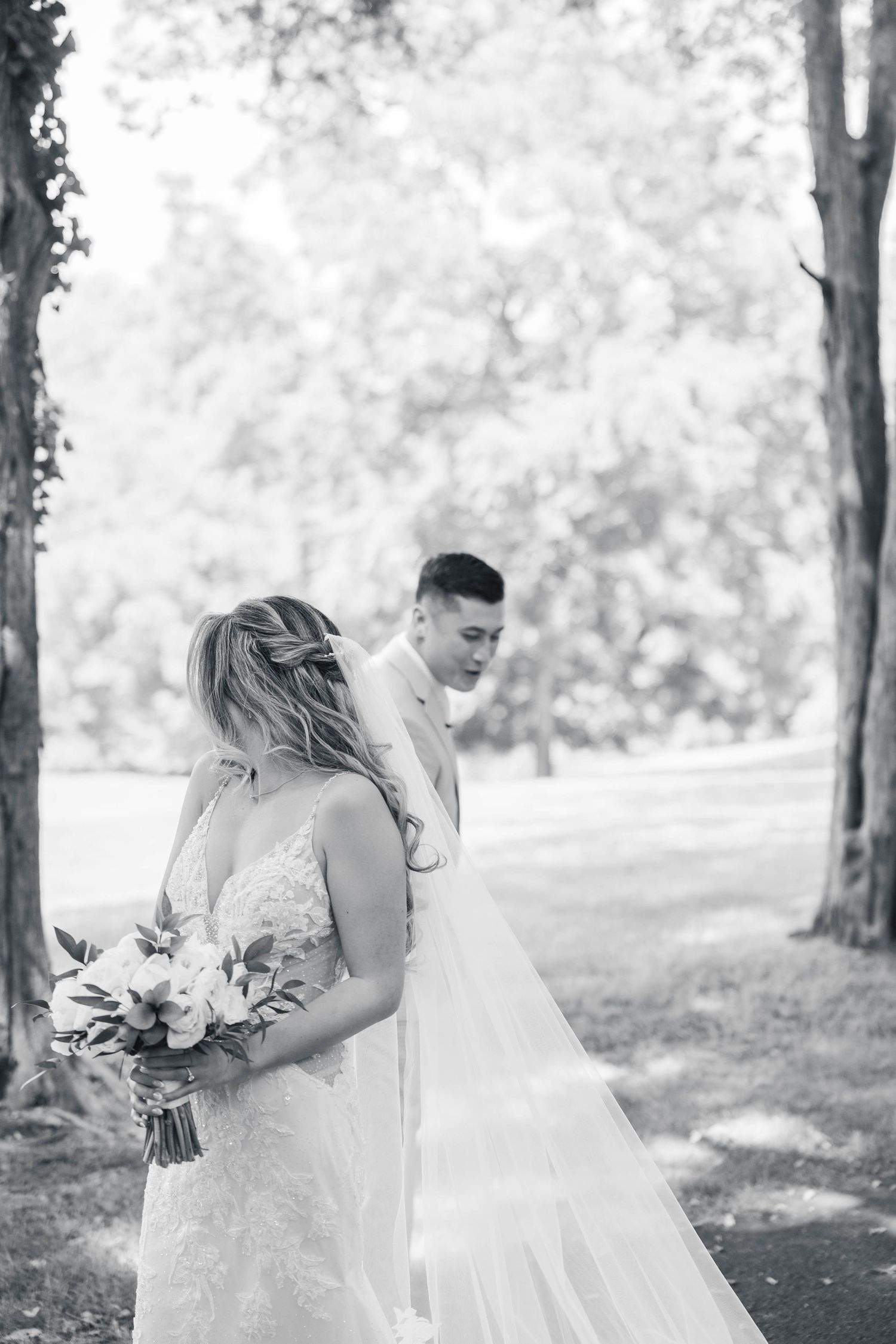 A bride in a flowing white dress holds a bouquet in a sun-dappled forest.