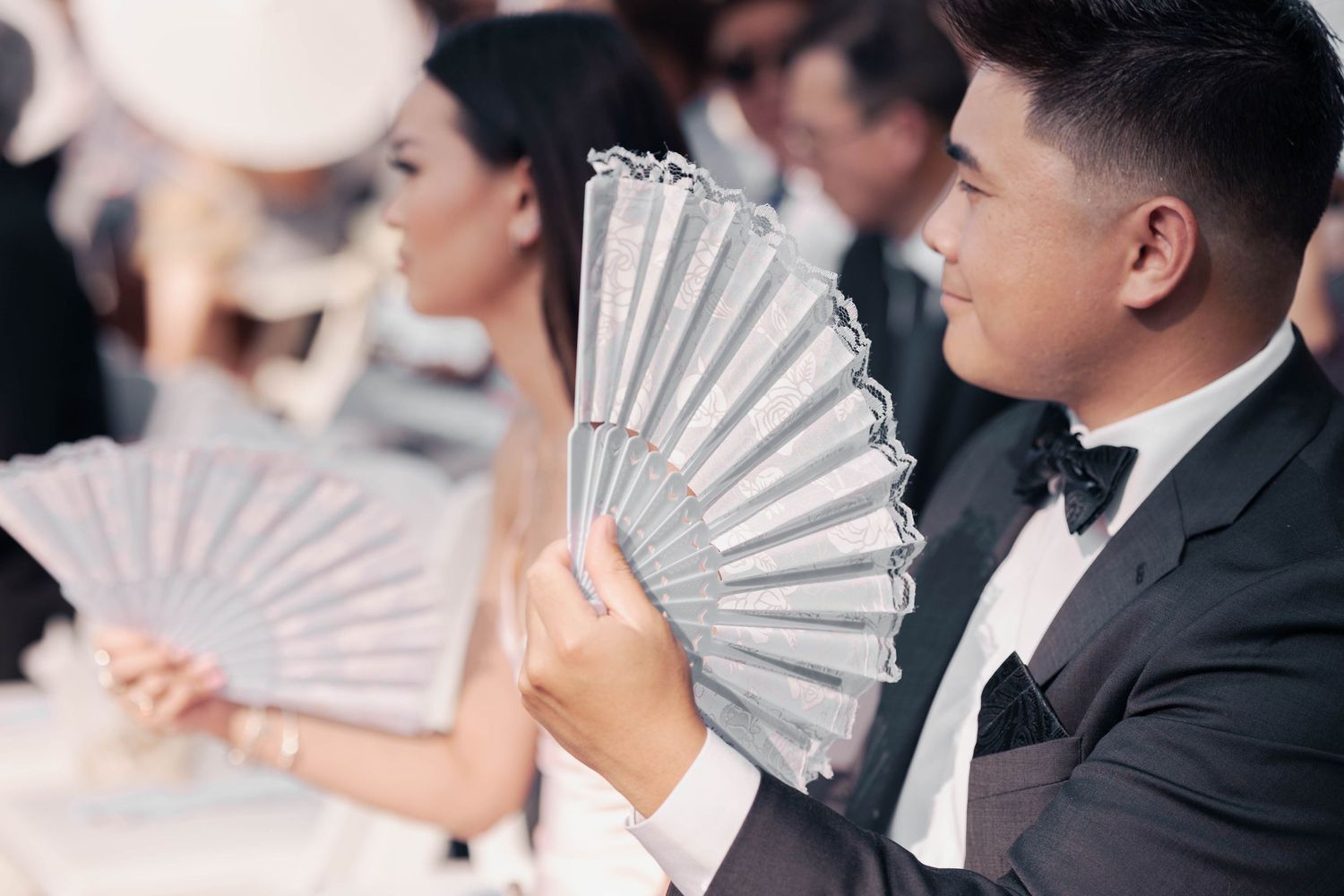 Wedding guests holding decorative white paper fans during outdoor ceremony.