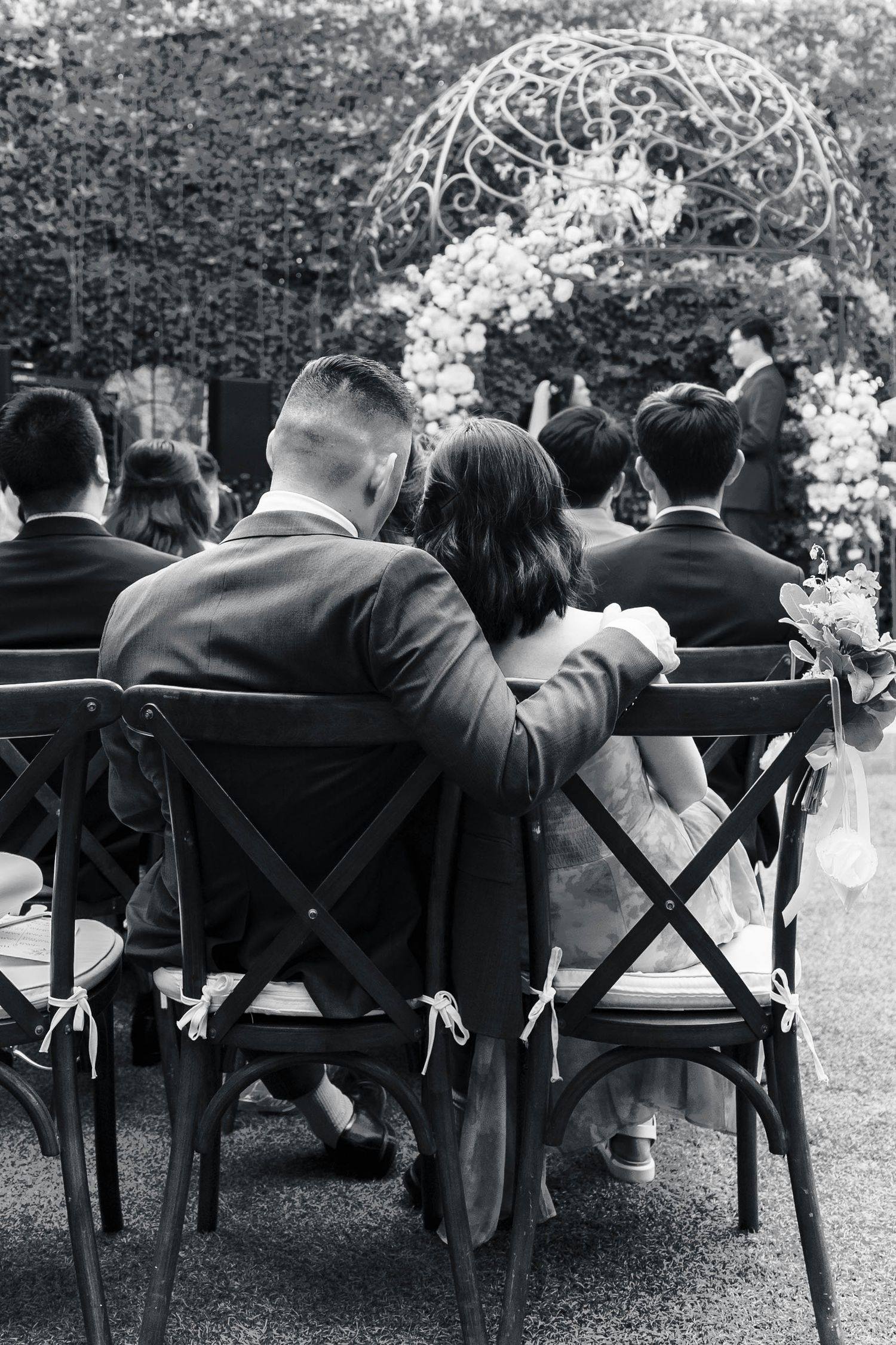 Wedding guests seated in wooden crossback chairs watch outdoor ceremony under floral arch in black and white.