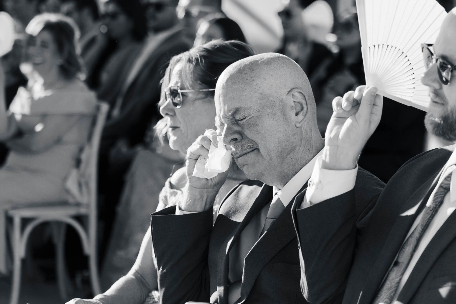 Elderly bald man in suit fans himself with hand fan at outdoor event, black and white photo.