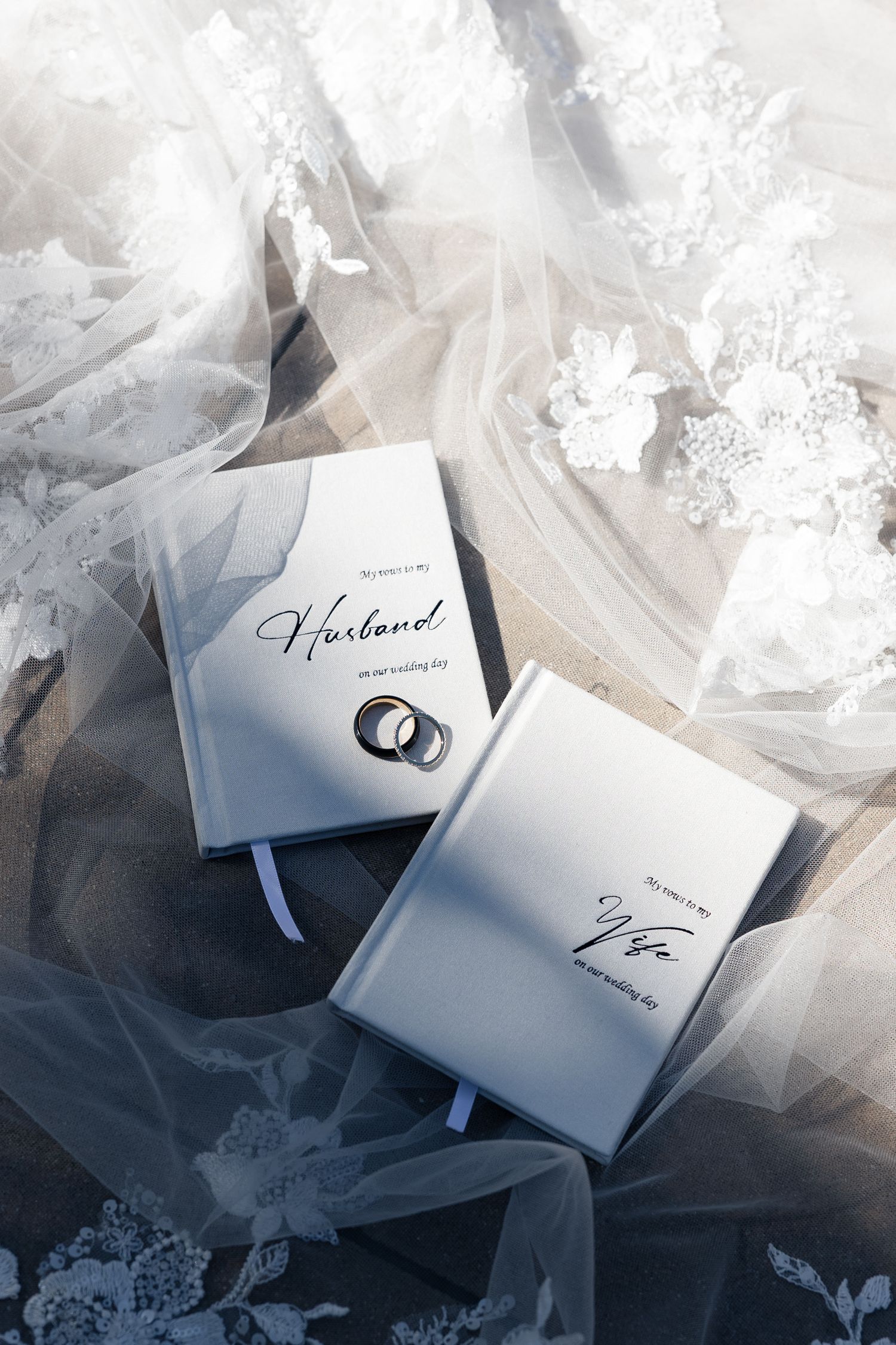Two wedding vow books labeled Husband and Wife with silver rings on a lace bridal veil backdrop.