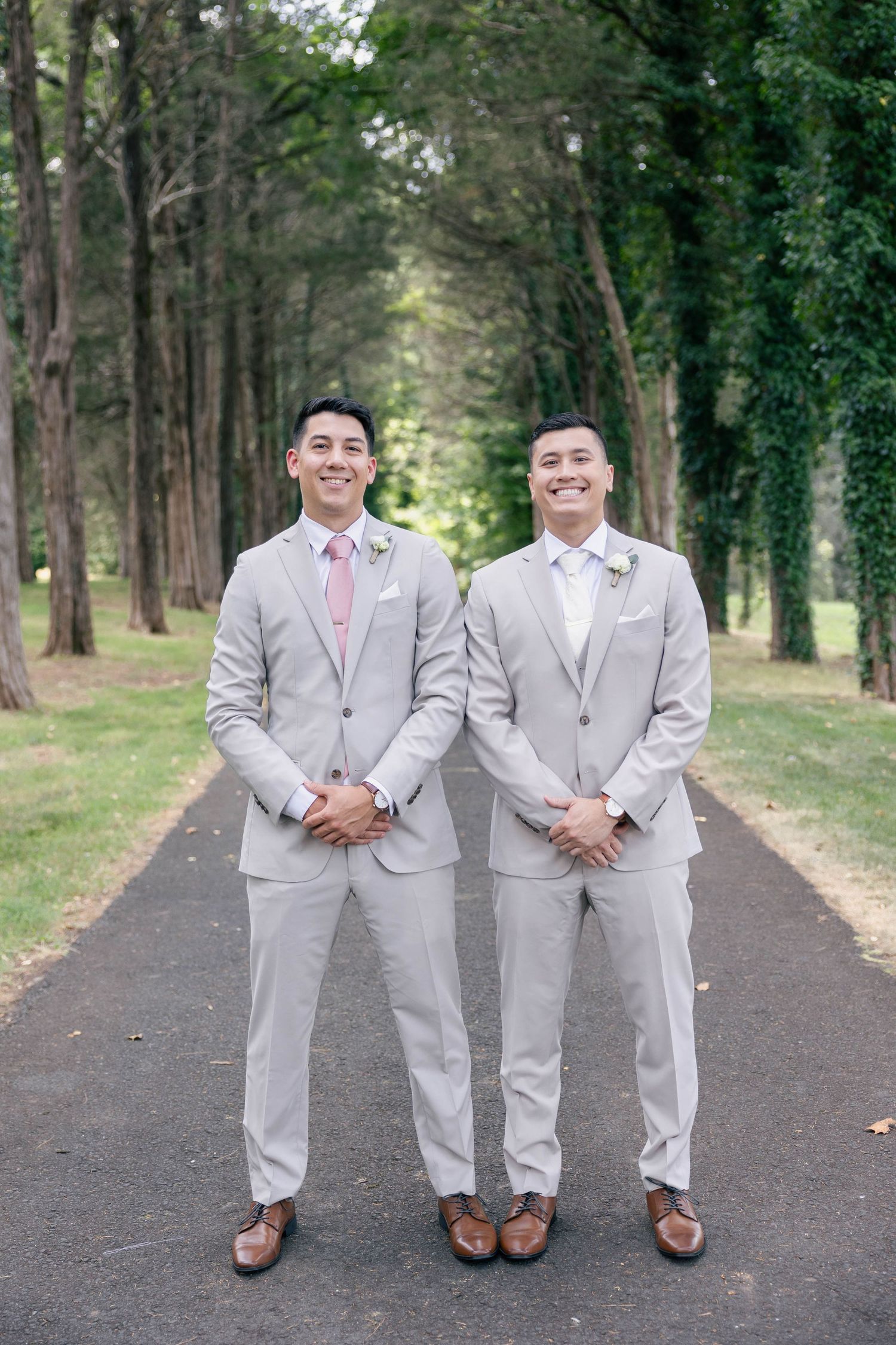 Two groomsmen in matching gray suits and pink ties pose on tree-lined trail.Groom and groomsmen wedding outfit inspiration in tan suit.