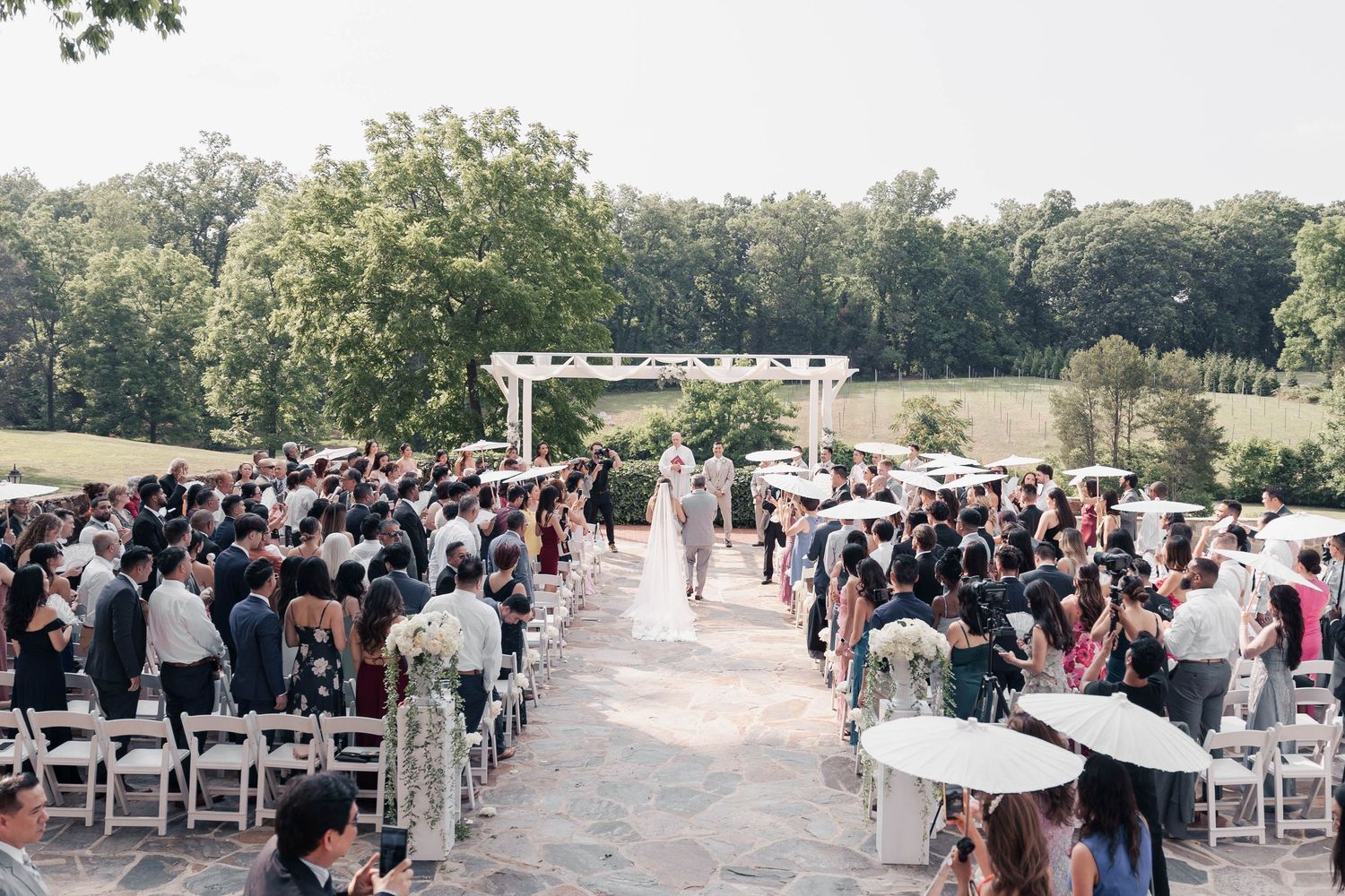 Wide shot of wedding guests seated in white chairs at outdoor venue.