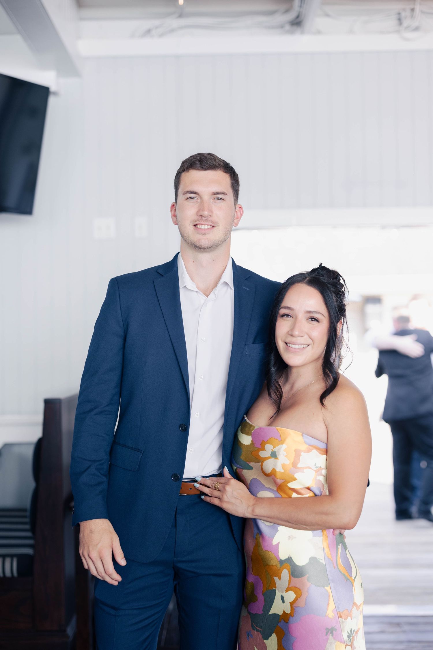 A couple in formal attire poses together at an event with navy suit and floral dress.