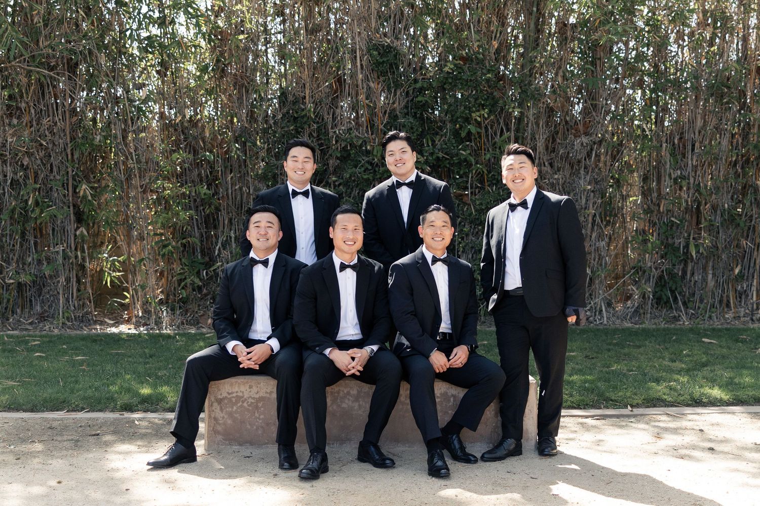 Six groomsmen in black tuxedos pose outdoors with bamboo trees, three seated and three standing behind them.