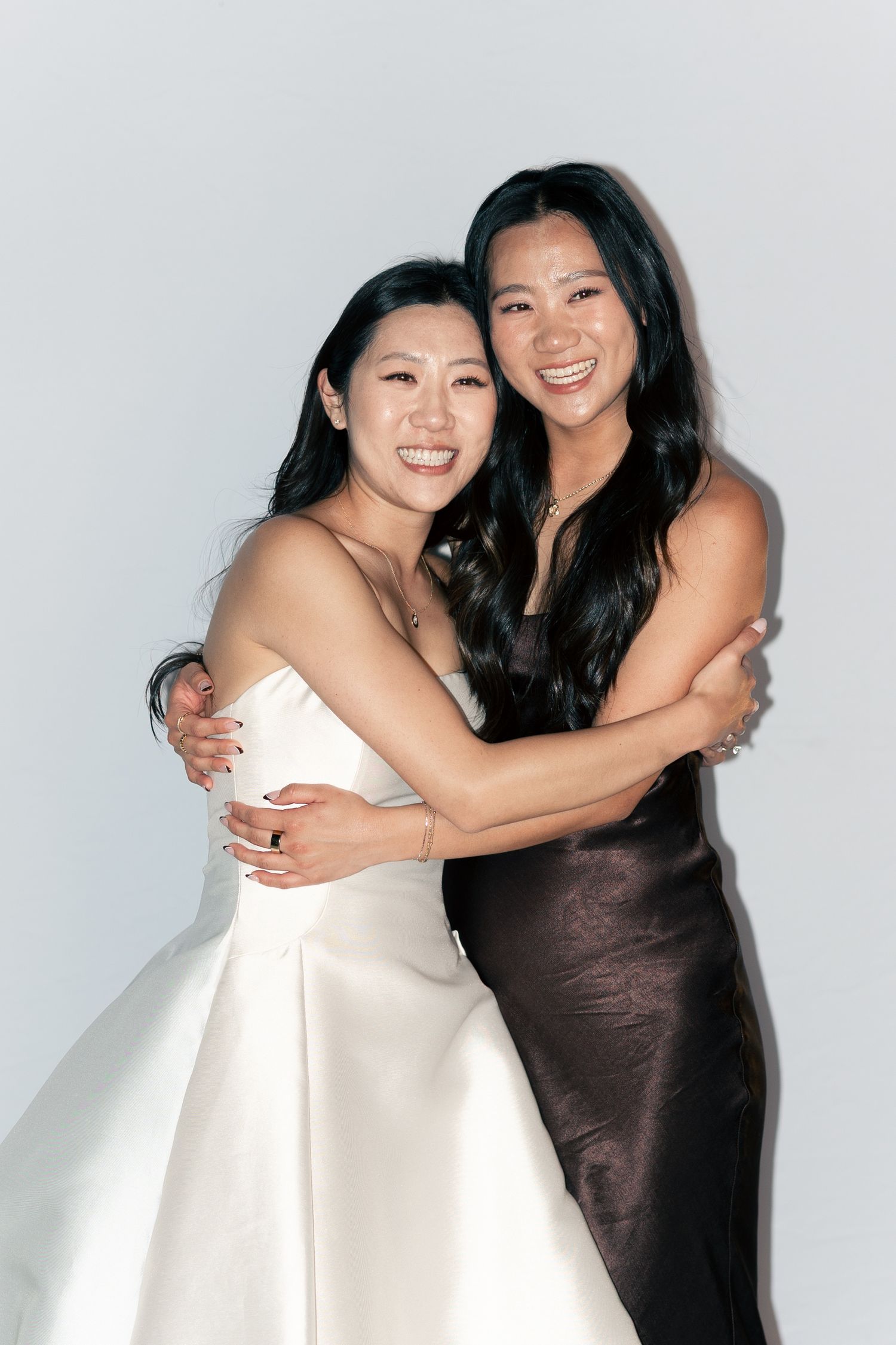 Two smiling women in elegant dresses, one in white bridal gown and one in brown satin, embracing warmly against a light background.
