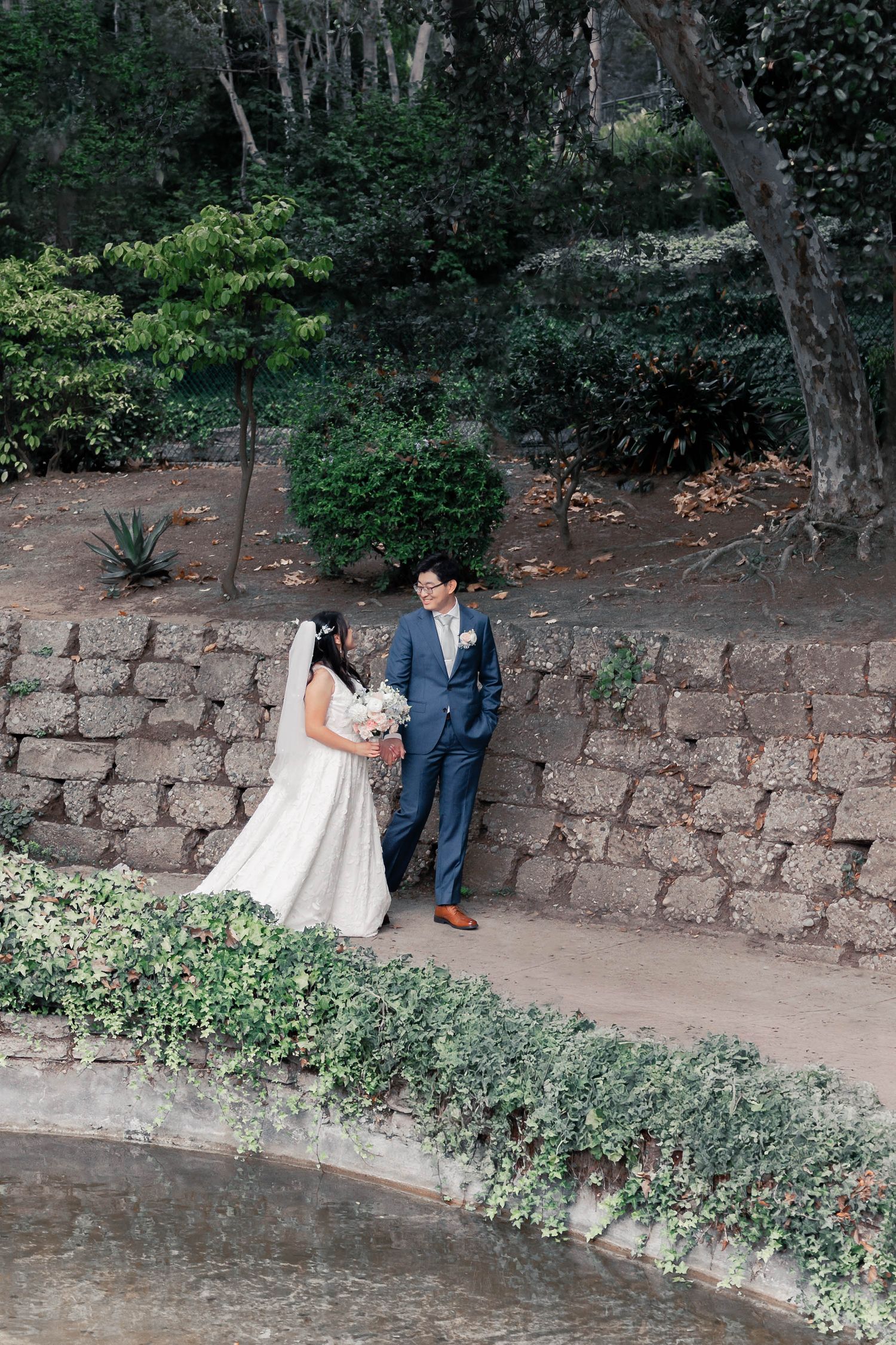 A couple in wedding attire sits together on a rustic stone wall surrounded by lush greenery in an outdoor garden setting.
