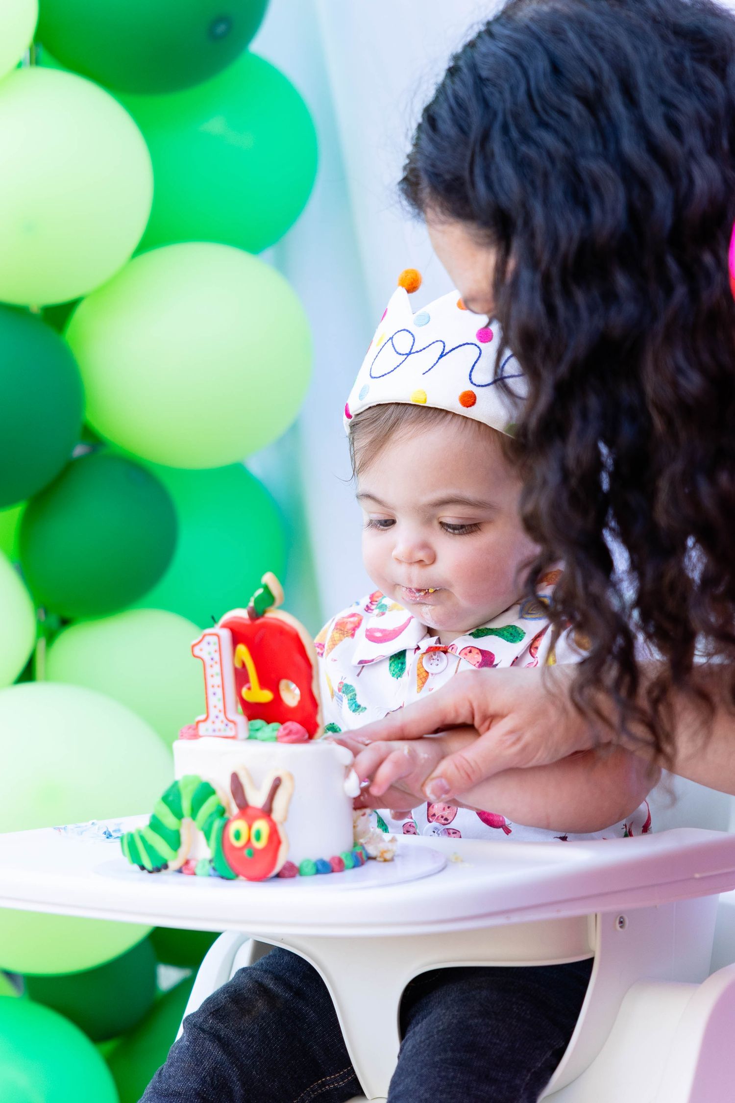 First birthday celebration with green balloon backdrop and decorated highchair for toddler wearing paper crown.