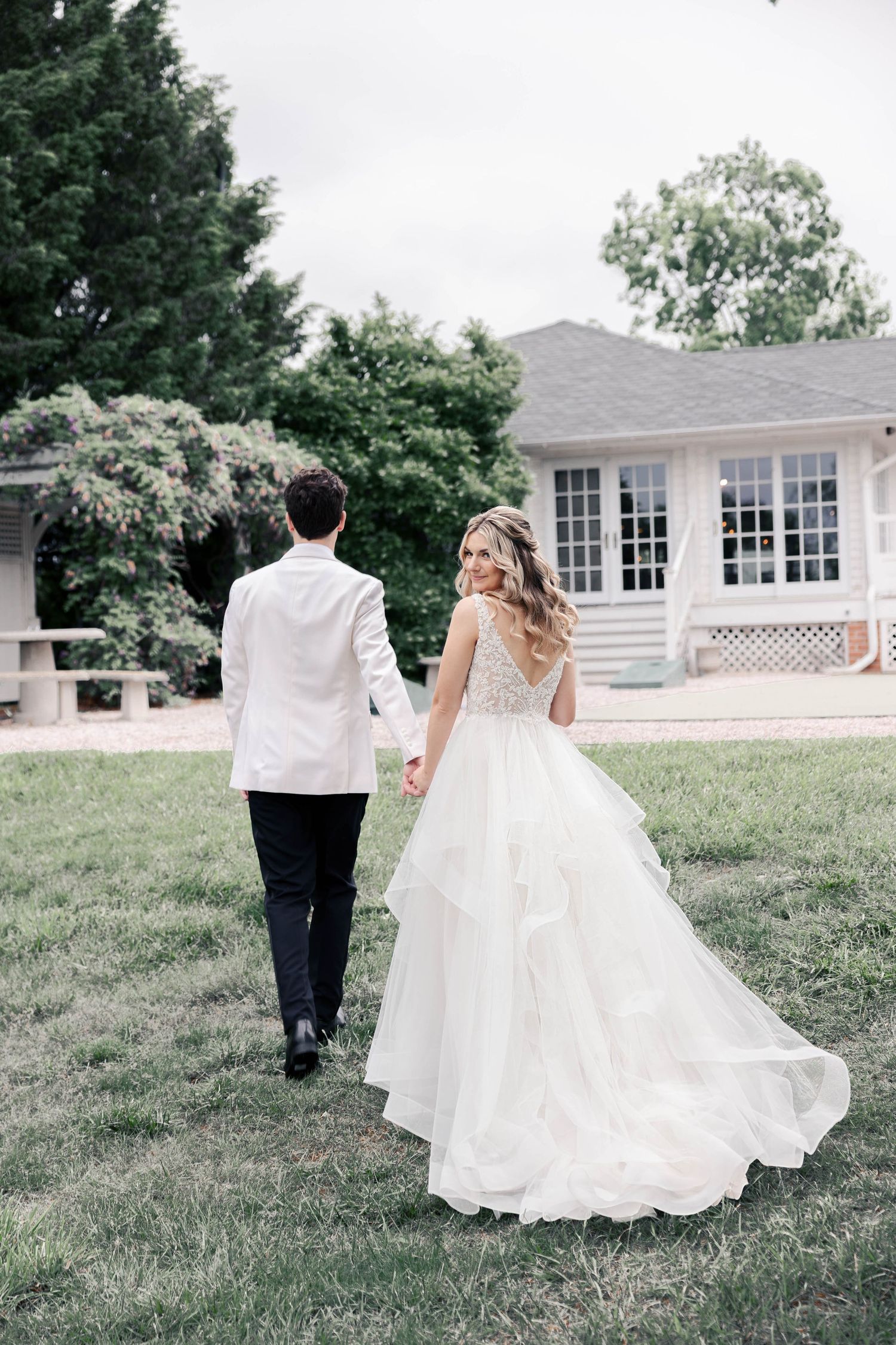 A couple in wedding attire walks hand in hand across a grassy lawn outside a white house with large windows.
