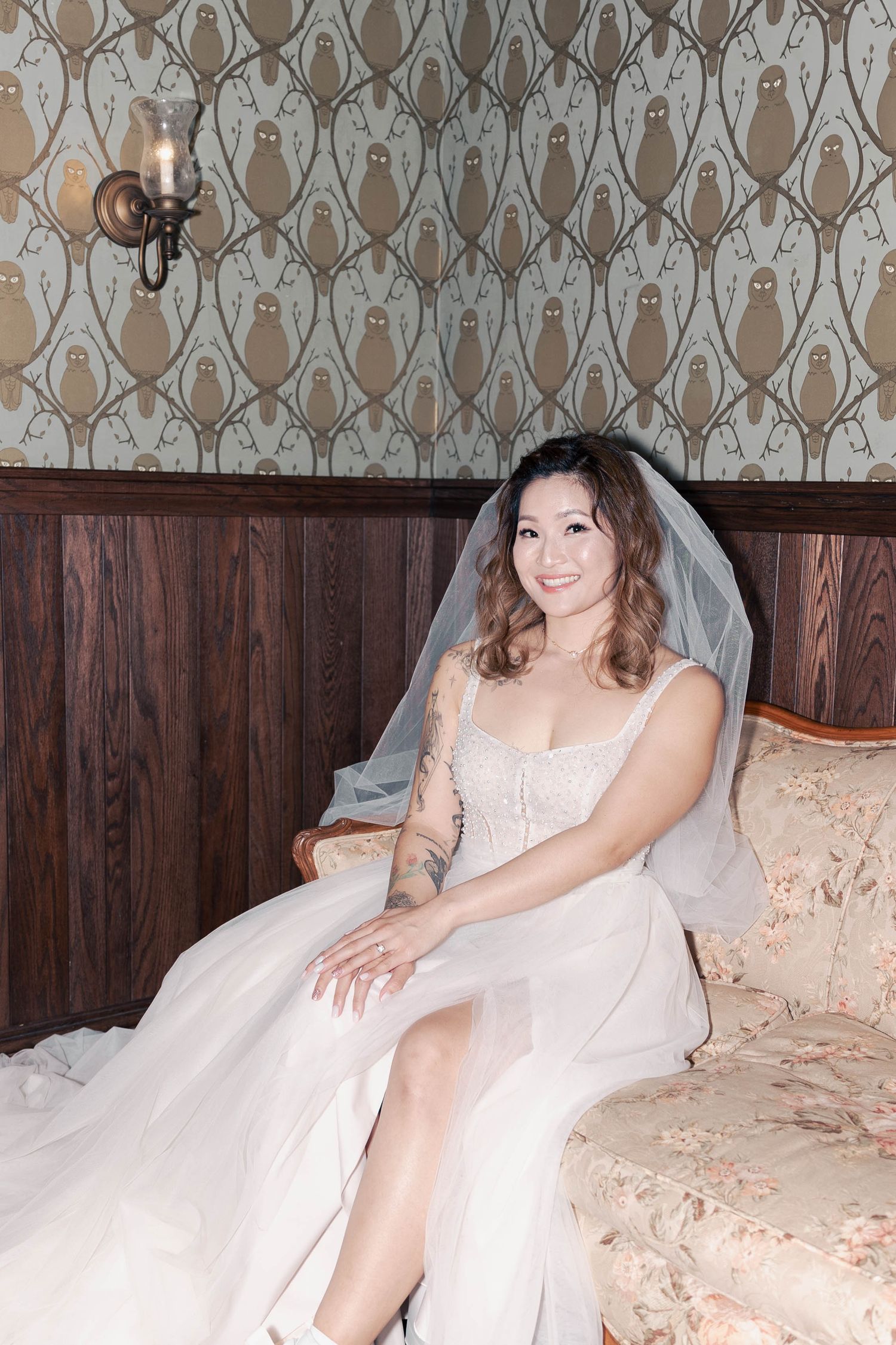 A bride in a white wedding dress and veil sits elegantly on a beige couch against a patterned wallpaper background.