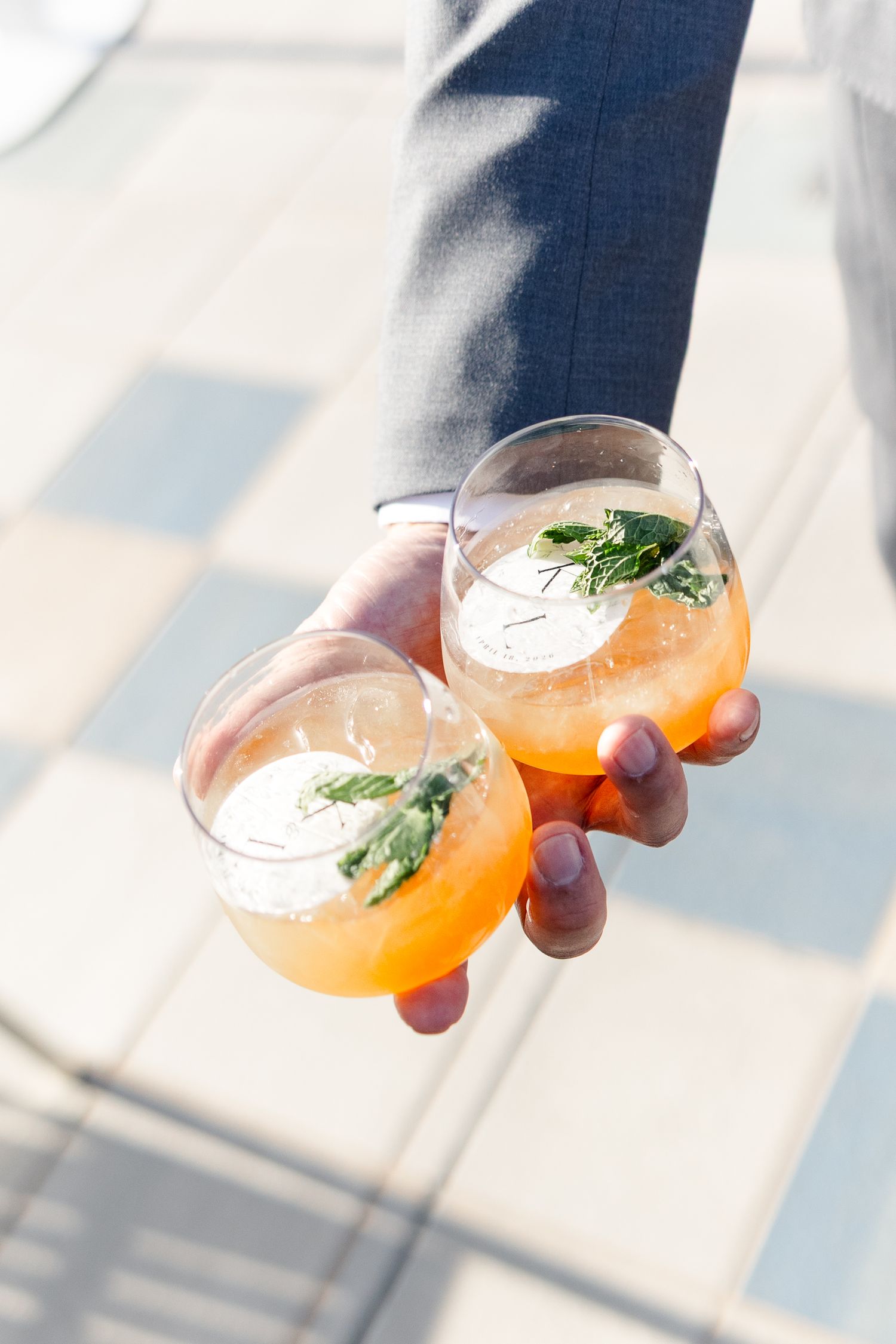 Hand holding two stemless glasses of orange cocktails garnished with fresh mint leaves on a sunny patio.