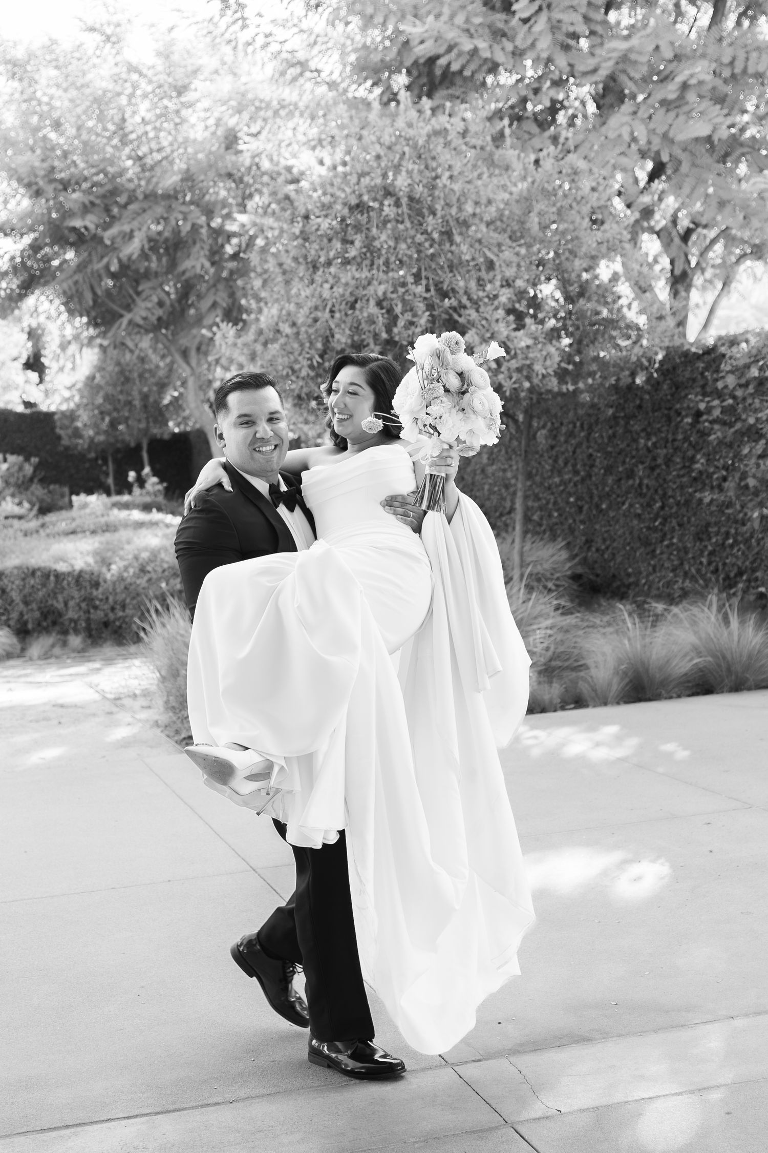Groom carries bride in white gown outdoors in black and white wedding photo with lush greenery backdrop.