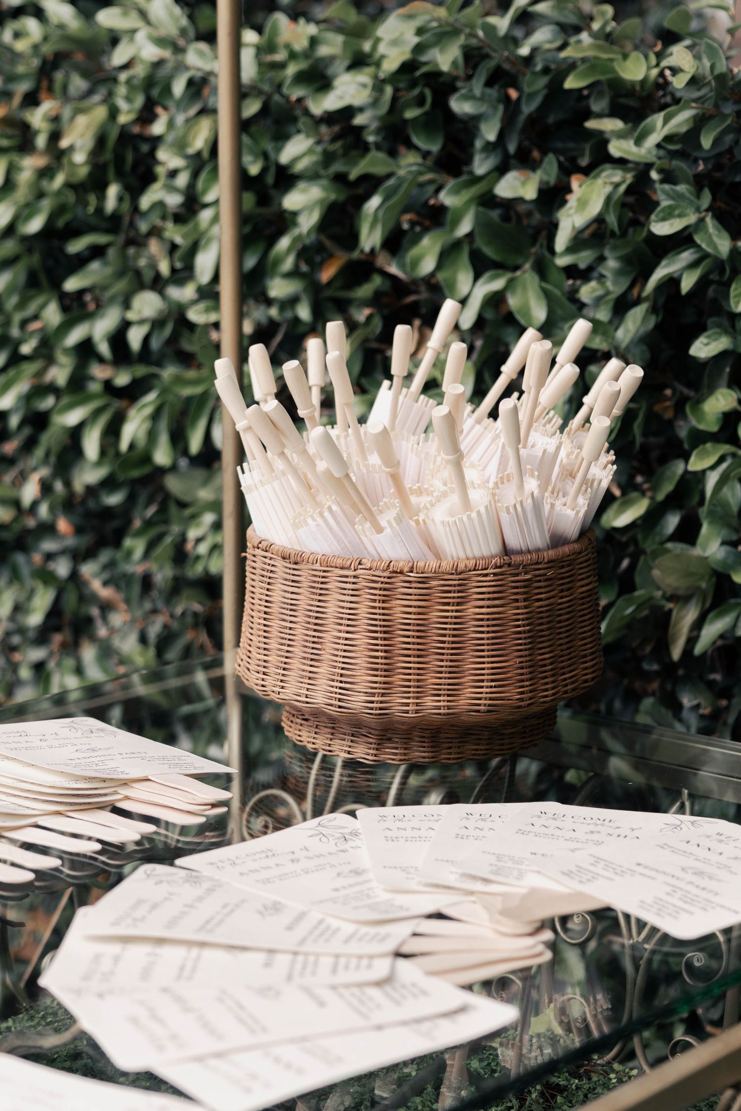 A woven basket filled with white paper straws and utensils sits on a table with scattered papers and foliage backdrop.