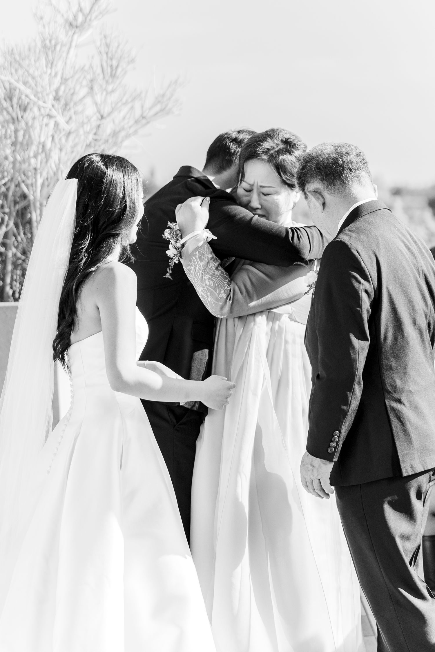 Black and white wedding photo of bride and groom embracing emotional family members during outdoor ceremony.
