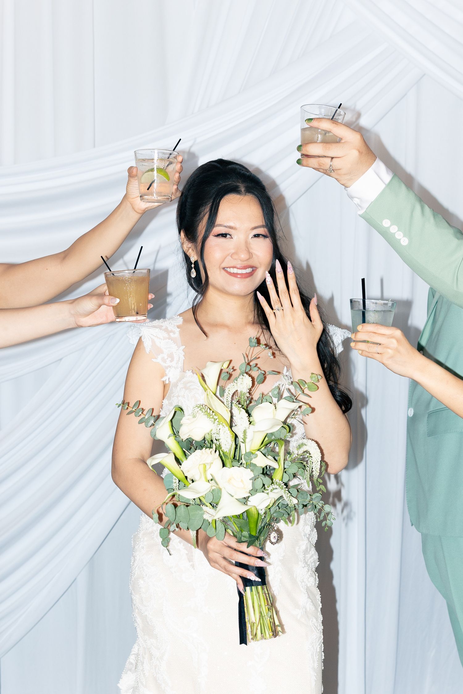 Smiling bride in lace gown holds white bouquet while guests raise cocktail glasses in a toast at elegant wedding reception.