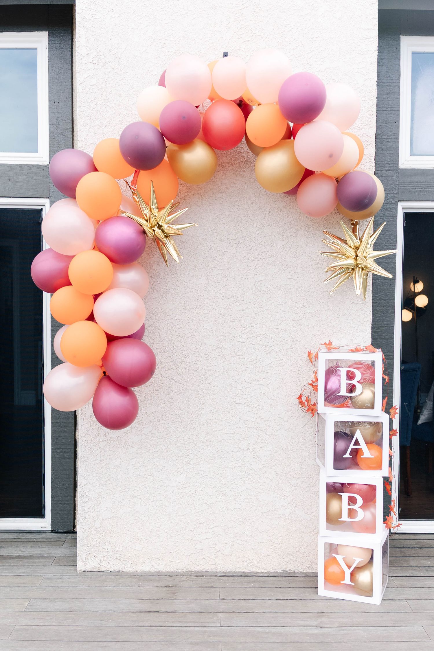 Decorative balloon arch in orange and purple shades with gold stars and white letter blocks against a light wall.