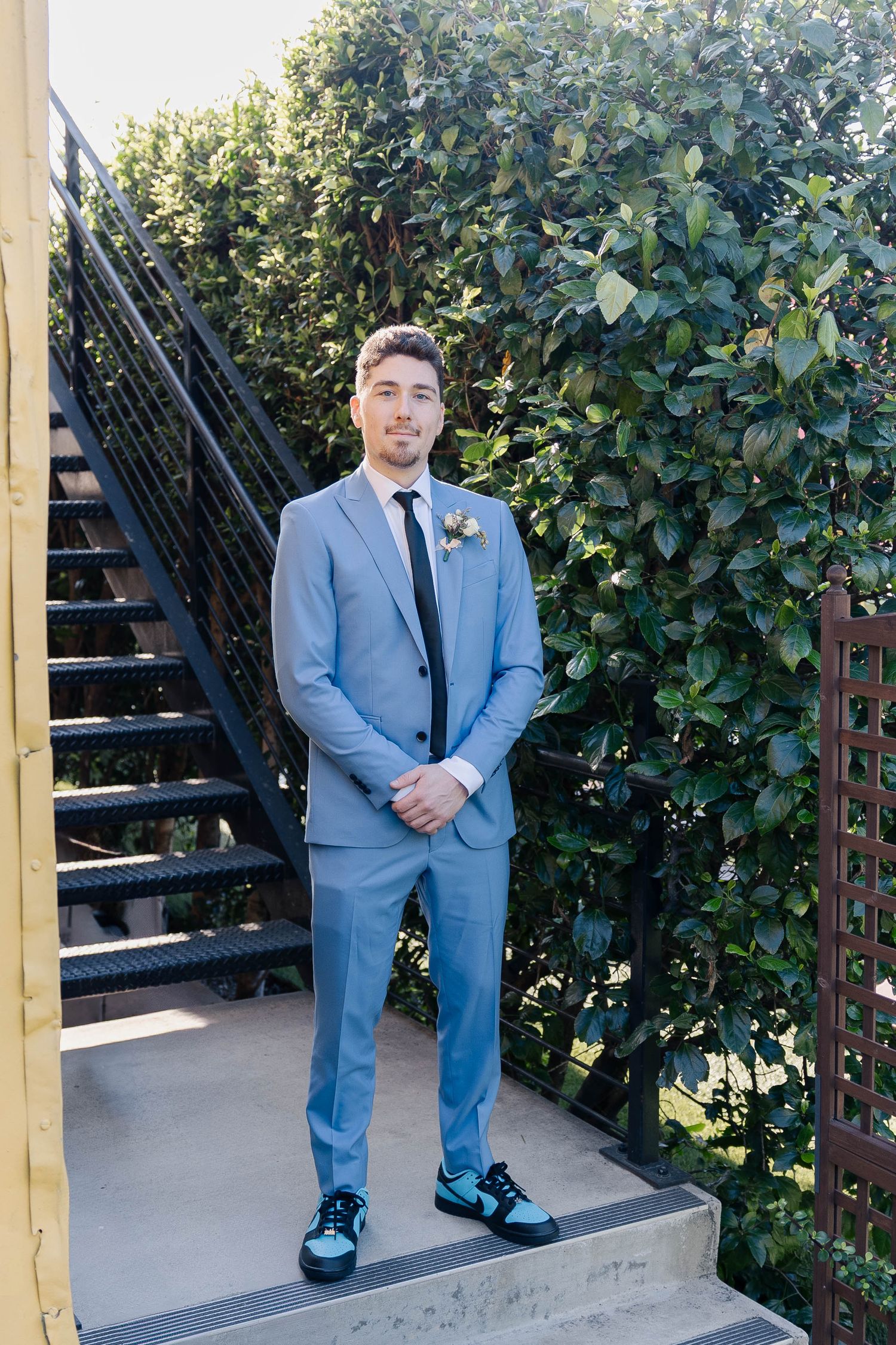 Groom in a powder blue suit poses next to greenery and stairs in outdoor wedding photos.