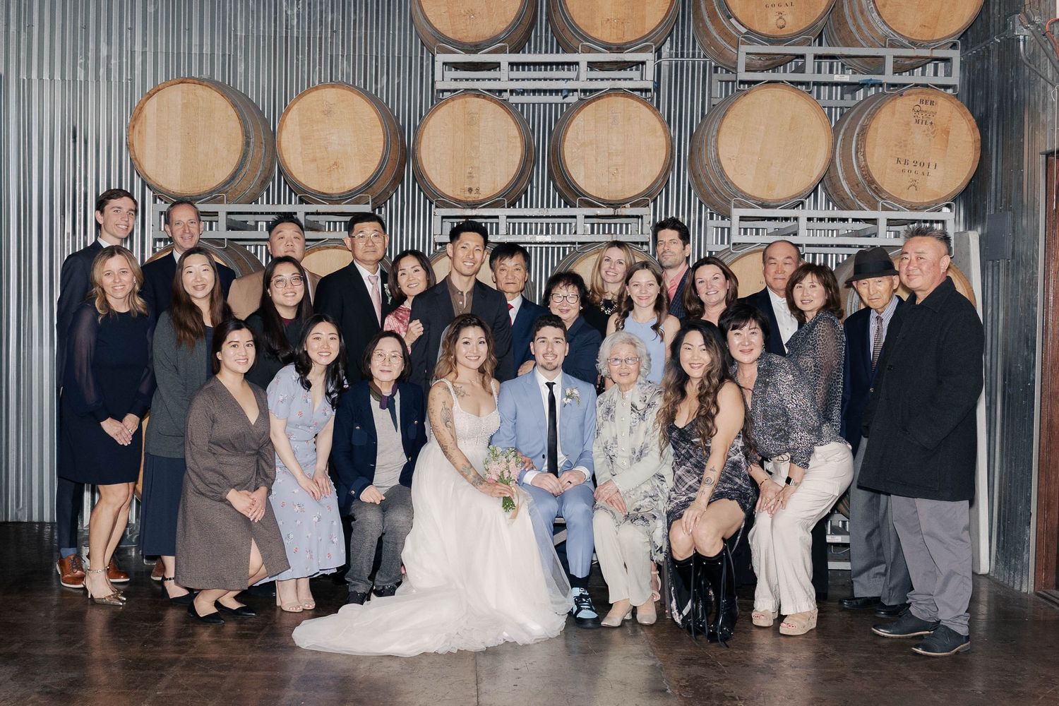 Large group photo in a rustic winery venue with wine barrels on wall and people gathered for a celebratory event.