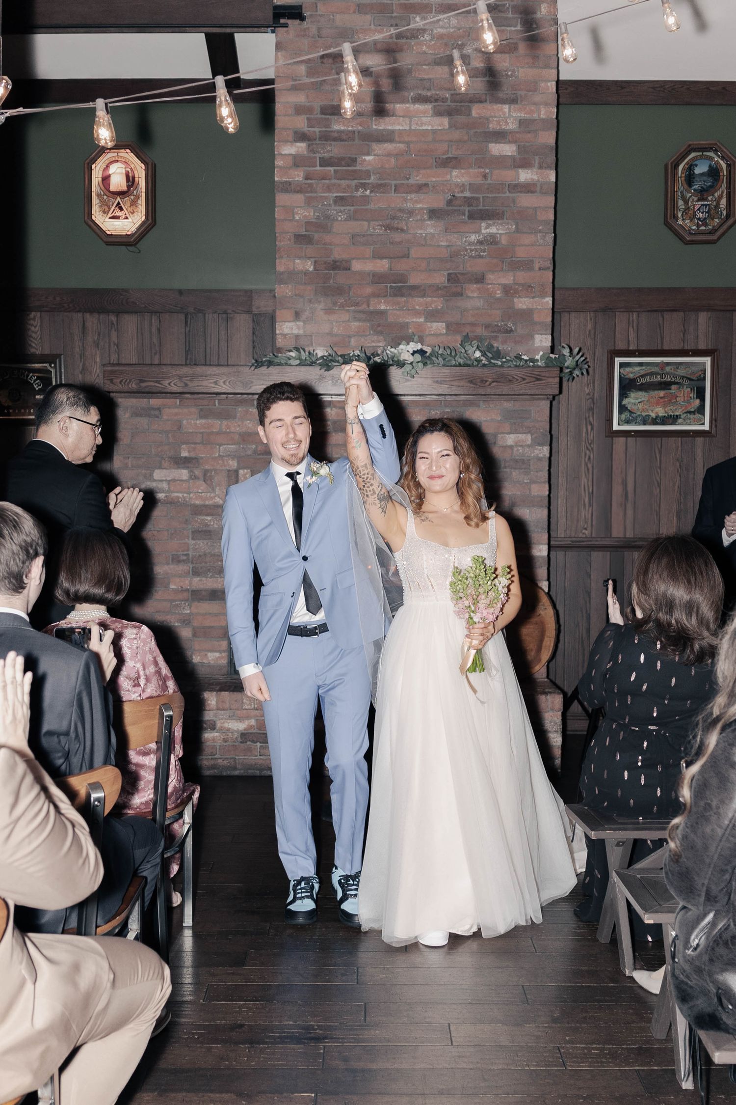 Wedding guests dance and celebrate on a wooden floor against brick walls with vintage lighting overhead.