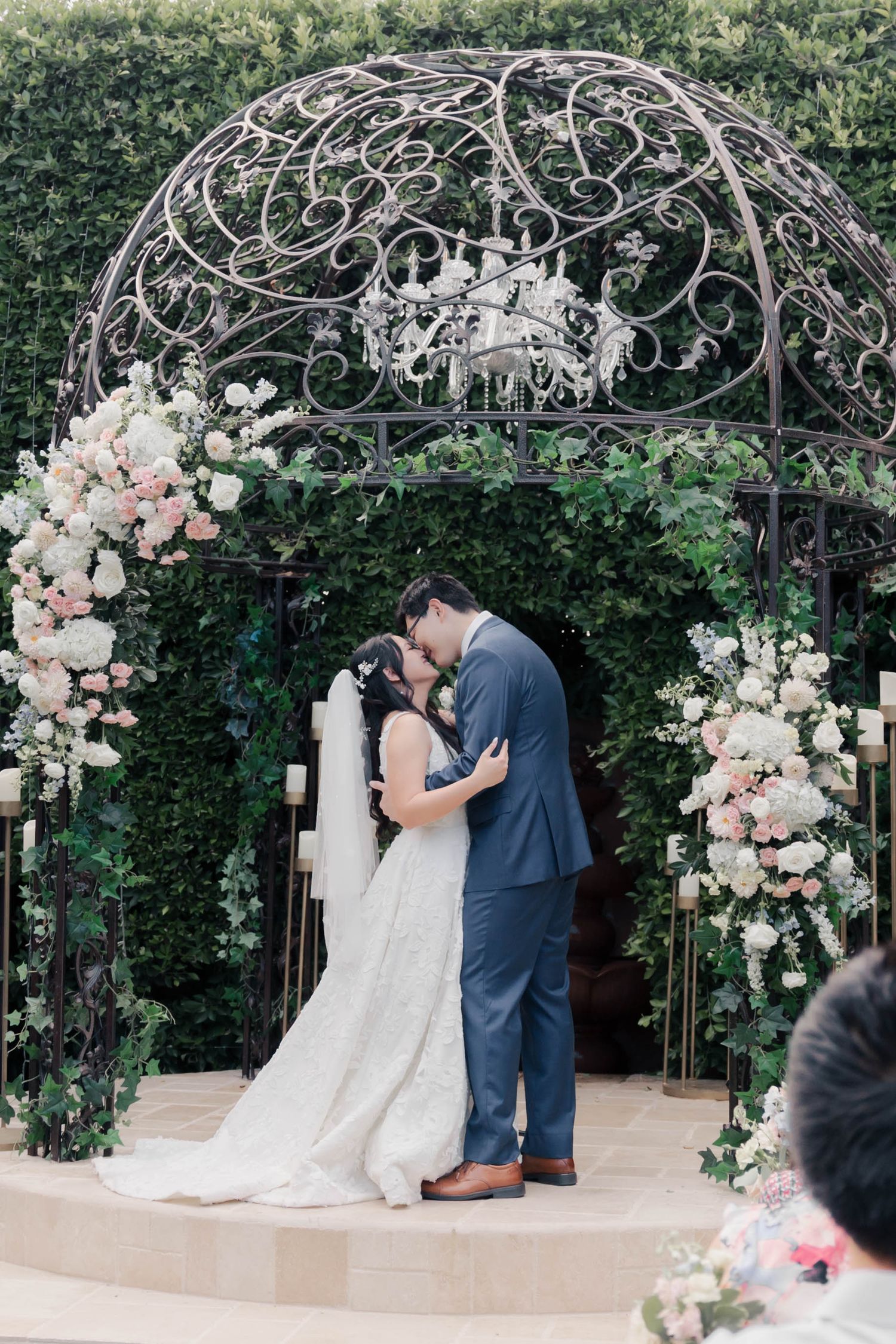 A romantic wedding ceremony moment under an ornate metal gazebo adorned with pink and white floral arrangements.