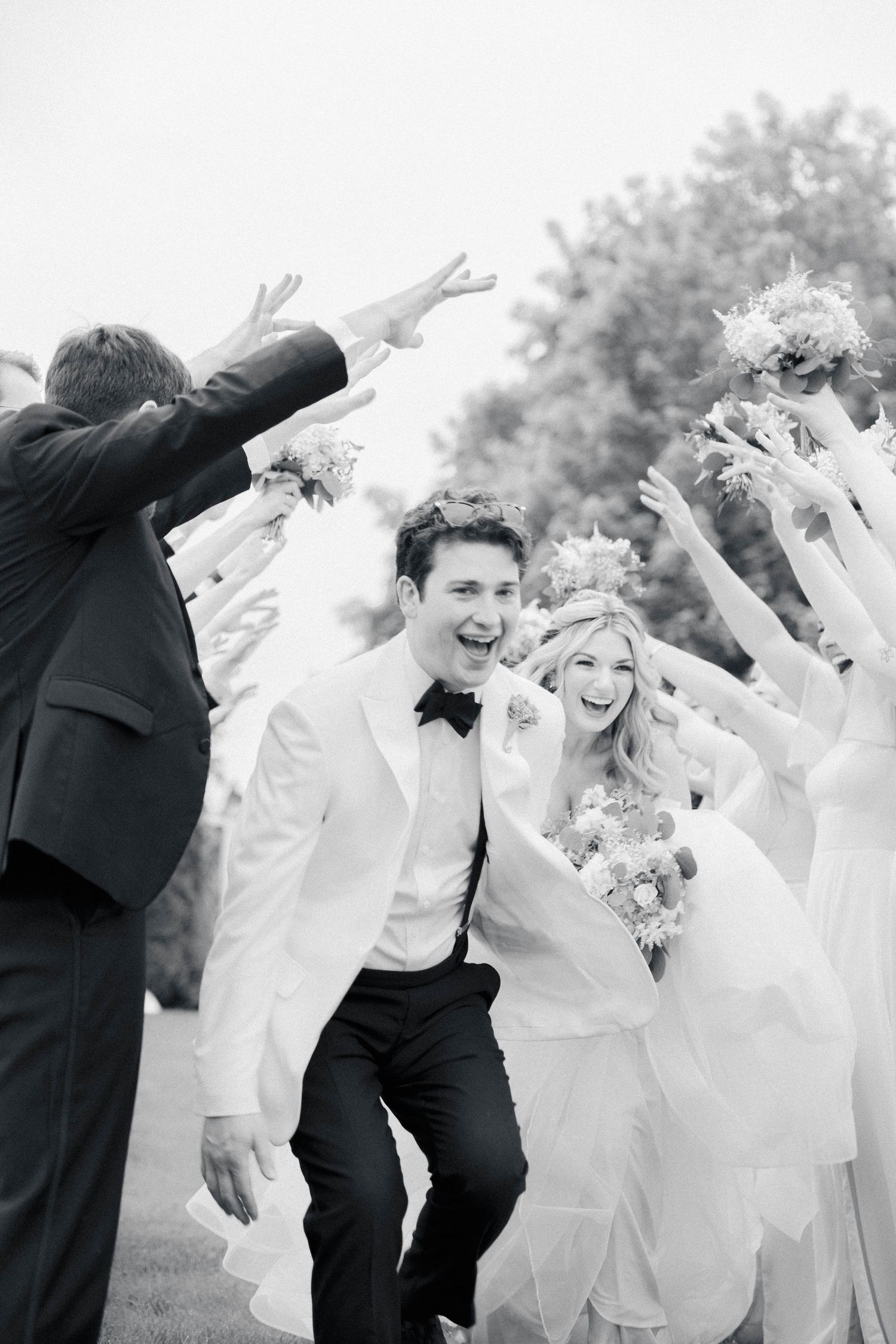 A joyful wedding party in black and white attire dances and celebrates together outdoors.