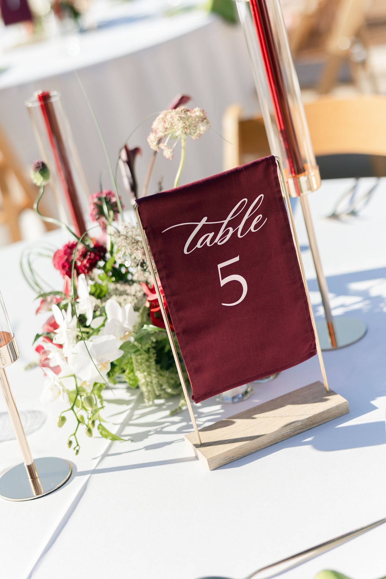 Elegant wedding table 5 marker with burgundy sign in gold frame, surrounded by floral centerpiece and red taper candles.