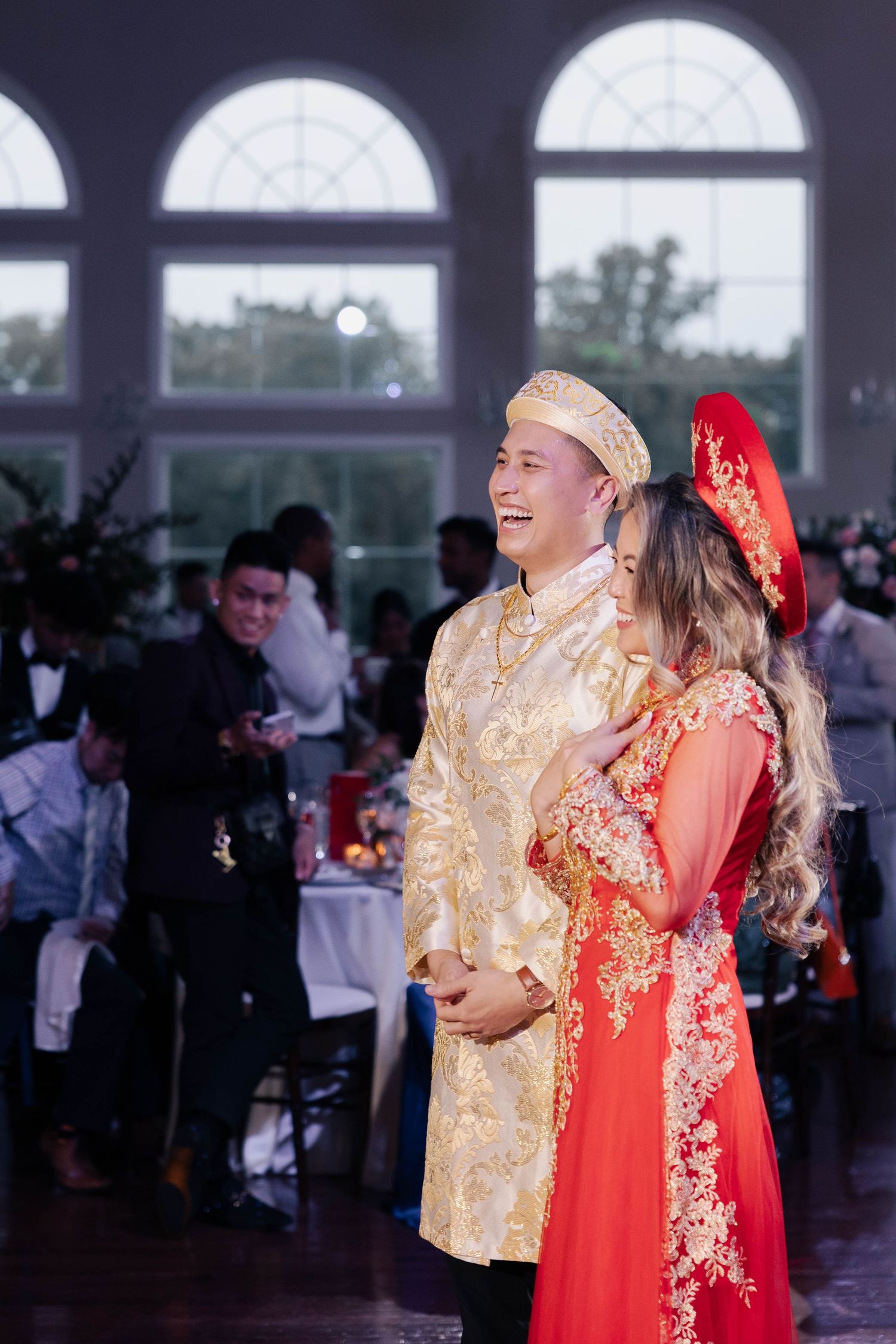 A couple in matching red and gold traditional Vietnamese attire laughing while dancing together at their wedding.